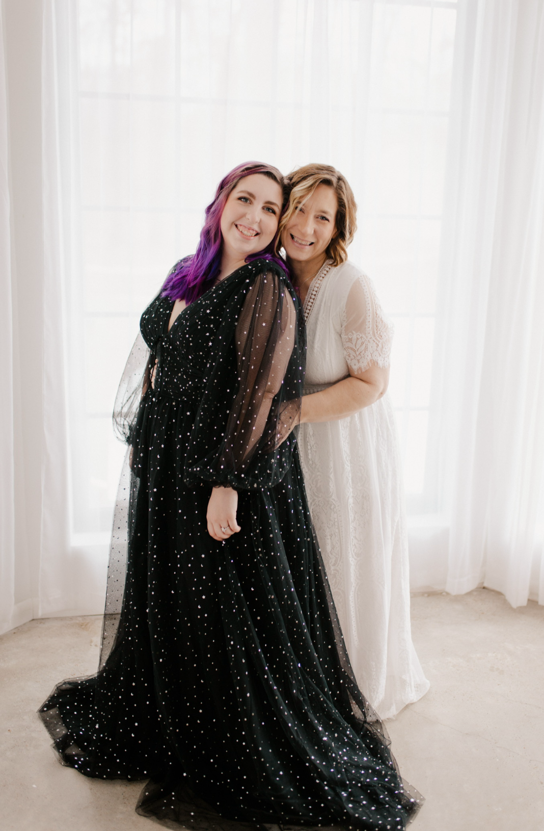 Two women standing together in front of sheer white curtains, smiling, with the younger woman wearing a black dress with sparkles and purple hair, and the older woman in a white lace dress.