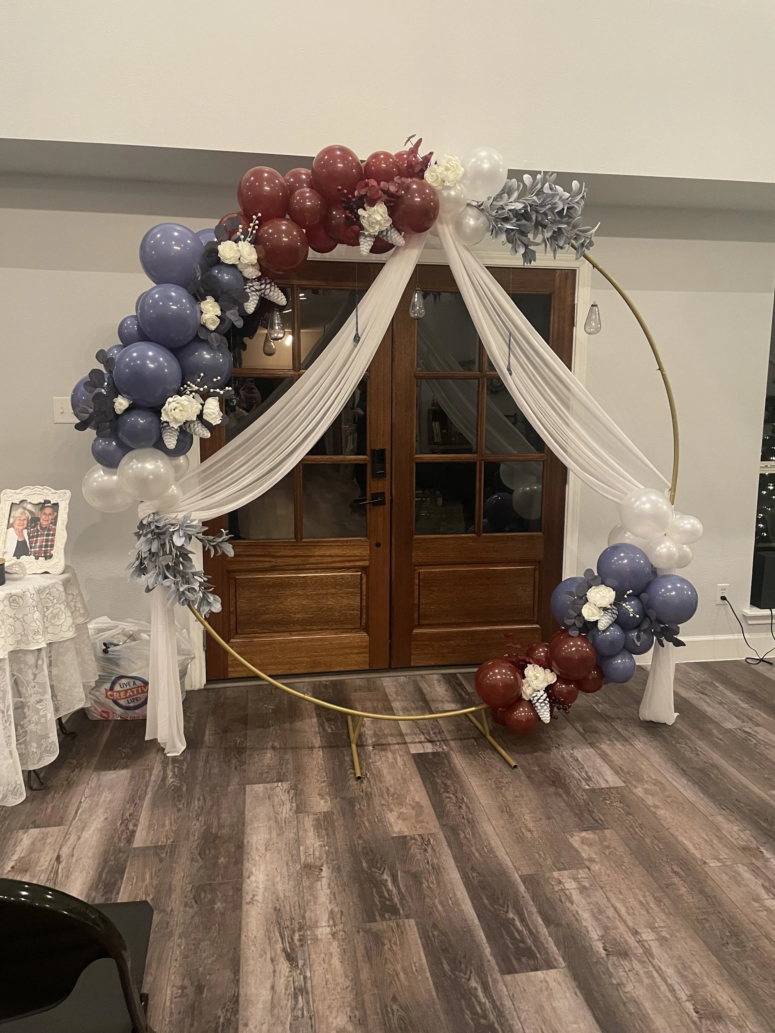A circular balloon arch with blue, white, and maroon balloons decorated with white flowers, gray leaves, and draped white fabric, set up indoors by a wooden door.