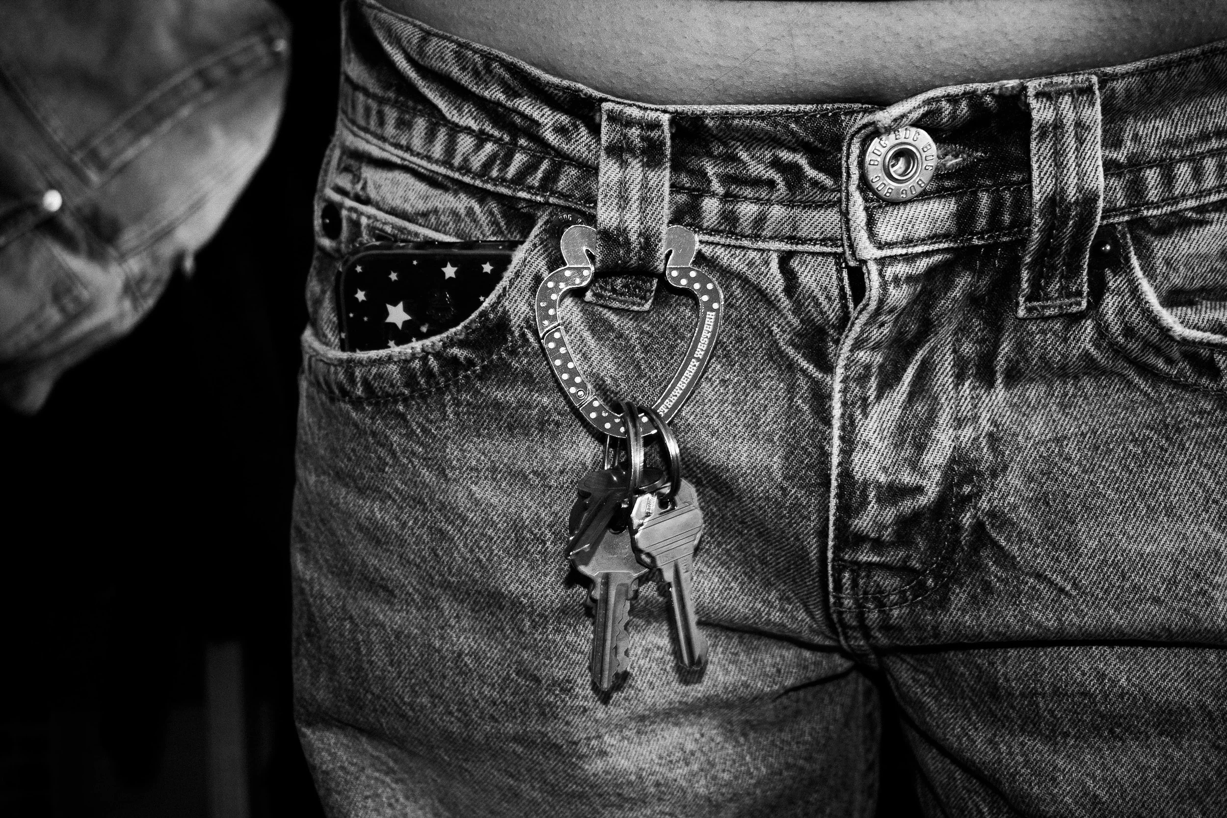 A person wearing jeans with a heart-shaped keychain clipped onto the belt loop, holding a set of keys, and a smartphone in the front pocket with a star pattern.
