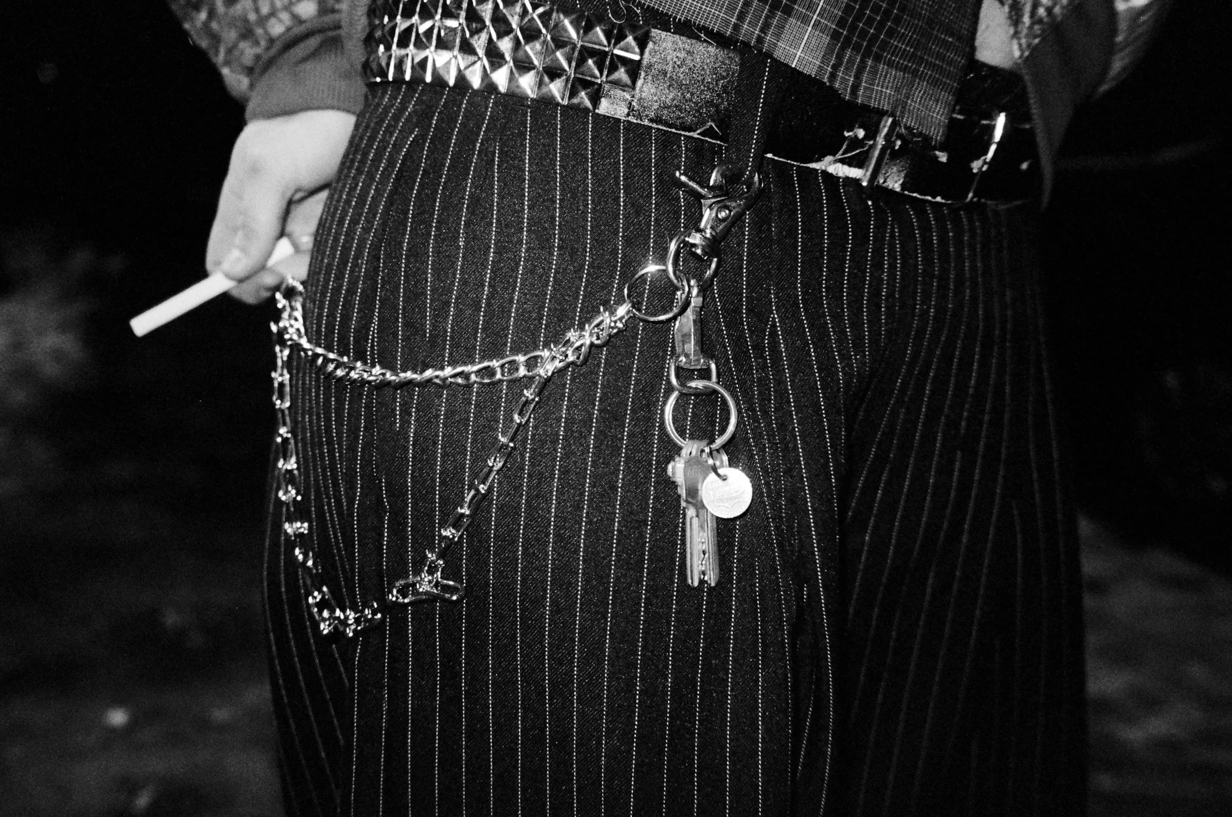 Close-up of a person's waist wearing striped pants and a checkered belt, with a chain hanging from the belt holding keys, and holding a cigarette.