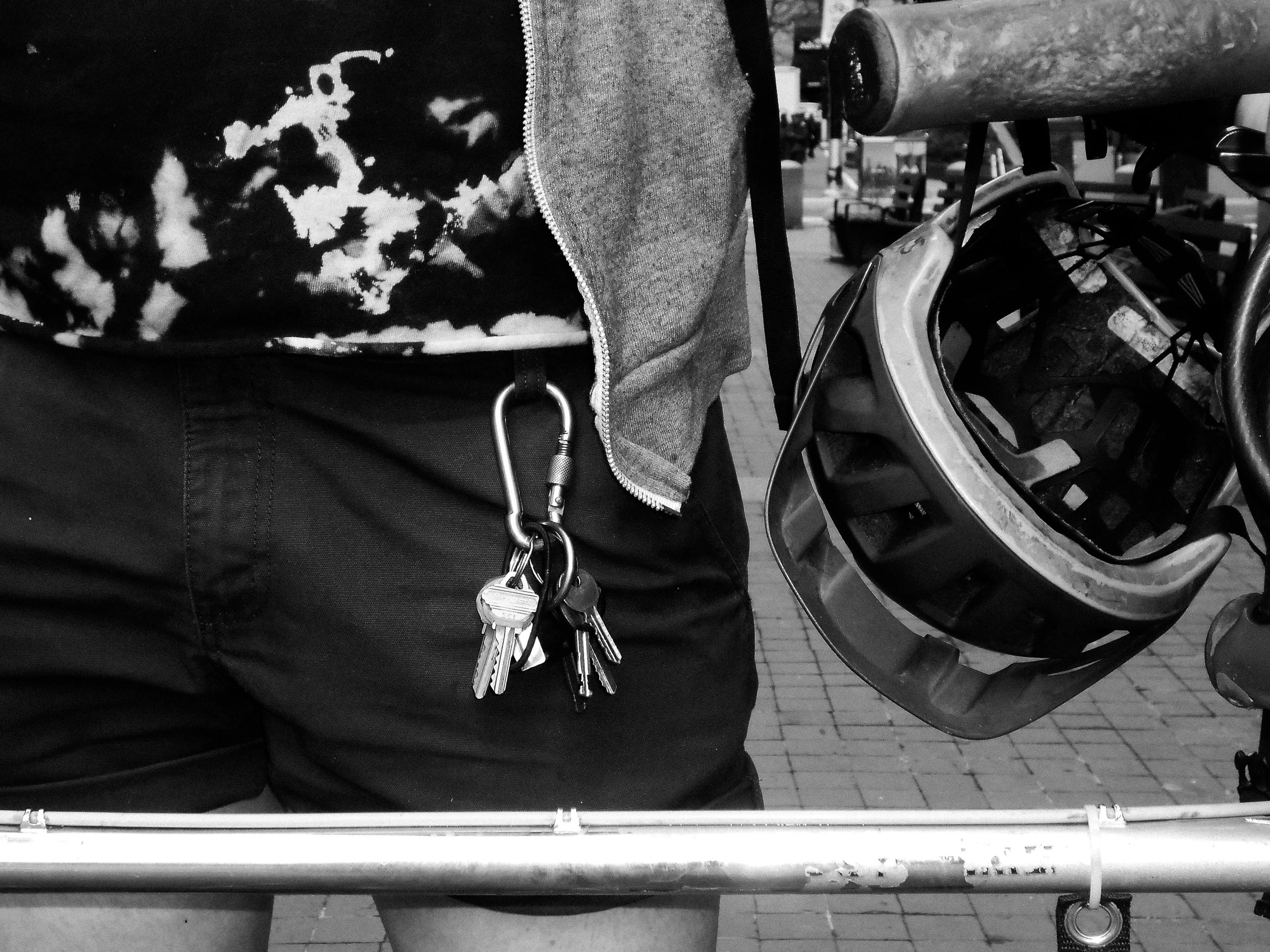 Close-up of a person wearing dark pants with keys attached to a carabiner clipped to the waist, and holding a bicycle helmet in front of a bike rack on a brick-paved sidewalk.