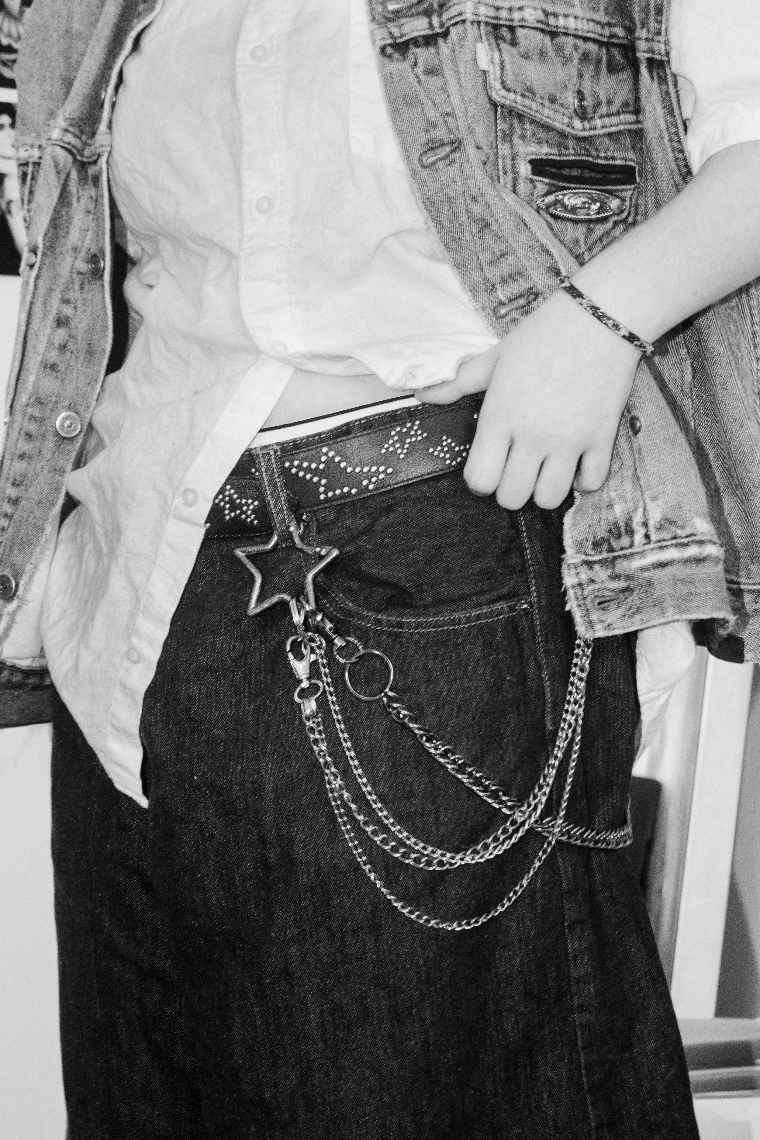 Close-up of a person wearing a white shirt, a denim vest, and dark jeans with a belt that has chains and a star-shaped buckle, along with chains and accessories hanging from the belt.