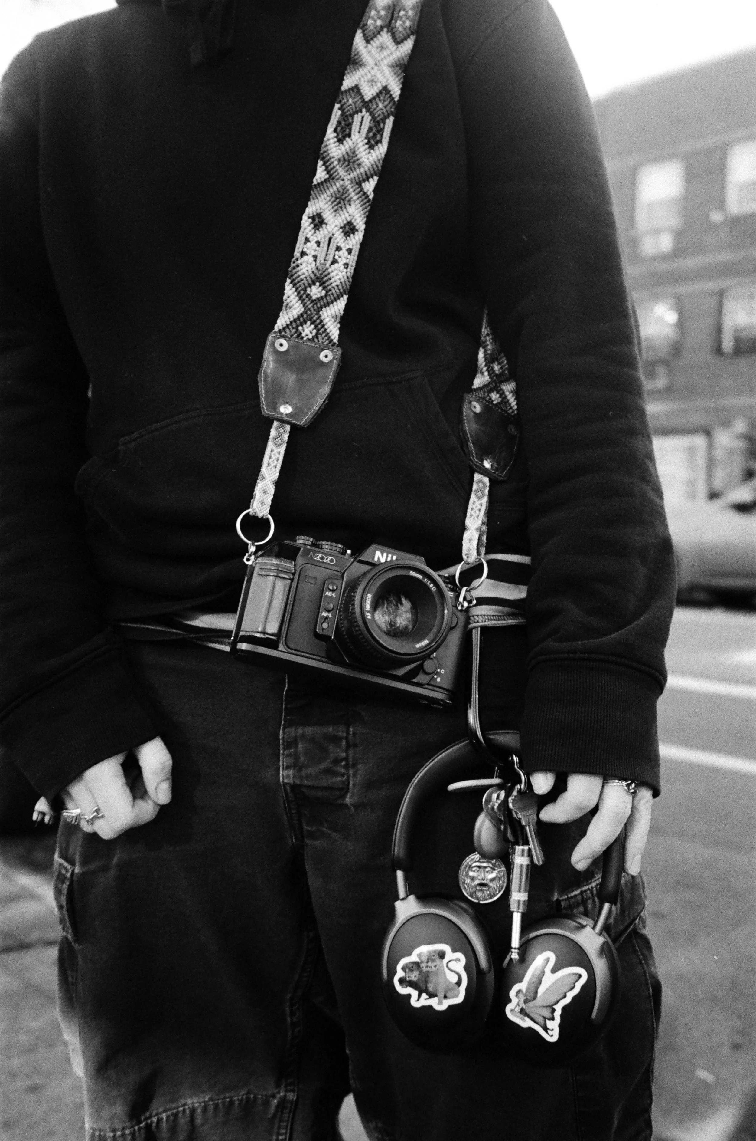 A person holding a set of headphones with stickers, wearing a camera around their neck, and dressed in dark clothing standing outdoors.