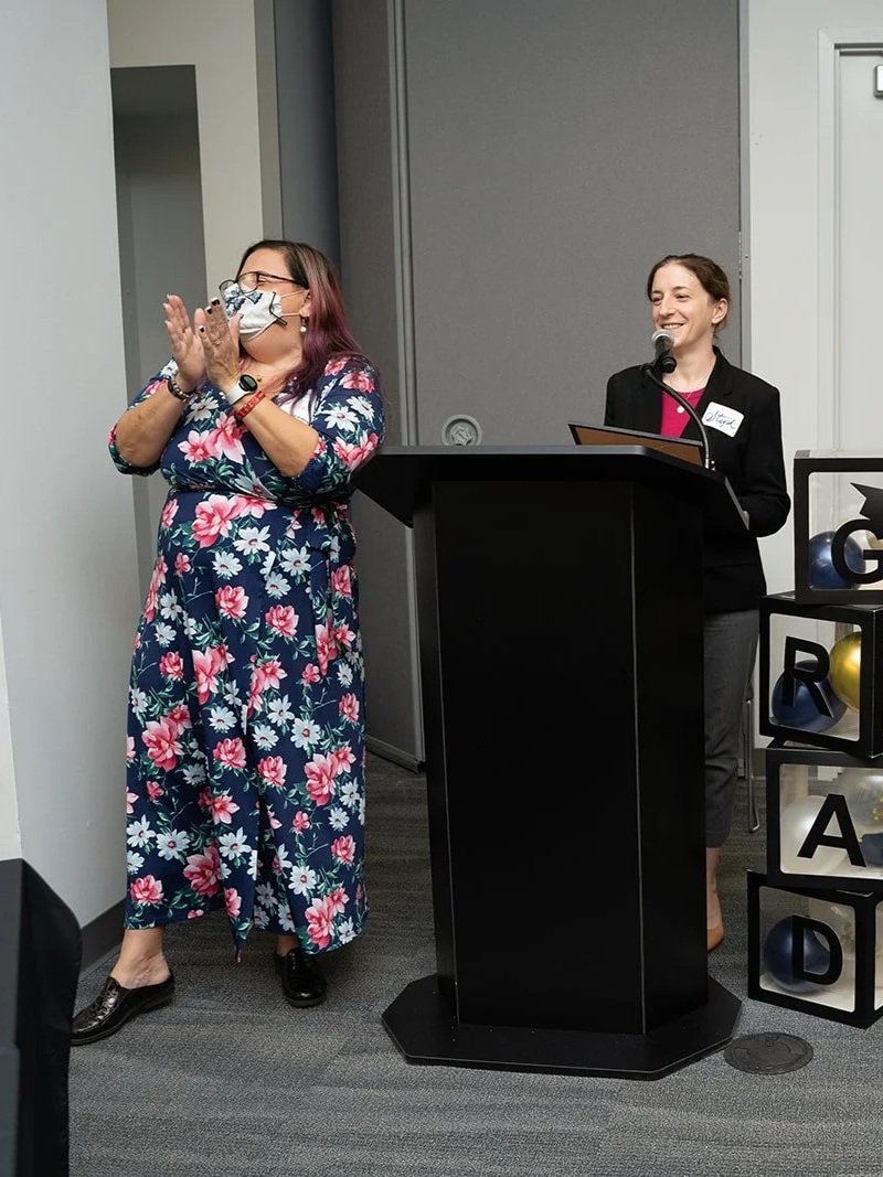 Two women standing at a podium, one clapping and the other speaking into a microphone, in an indoor setting with gray walls and a black carpet.