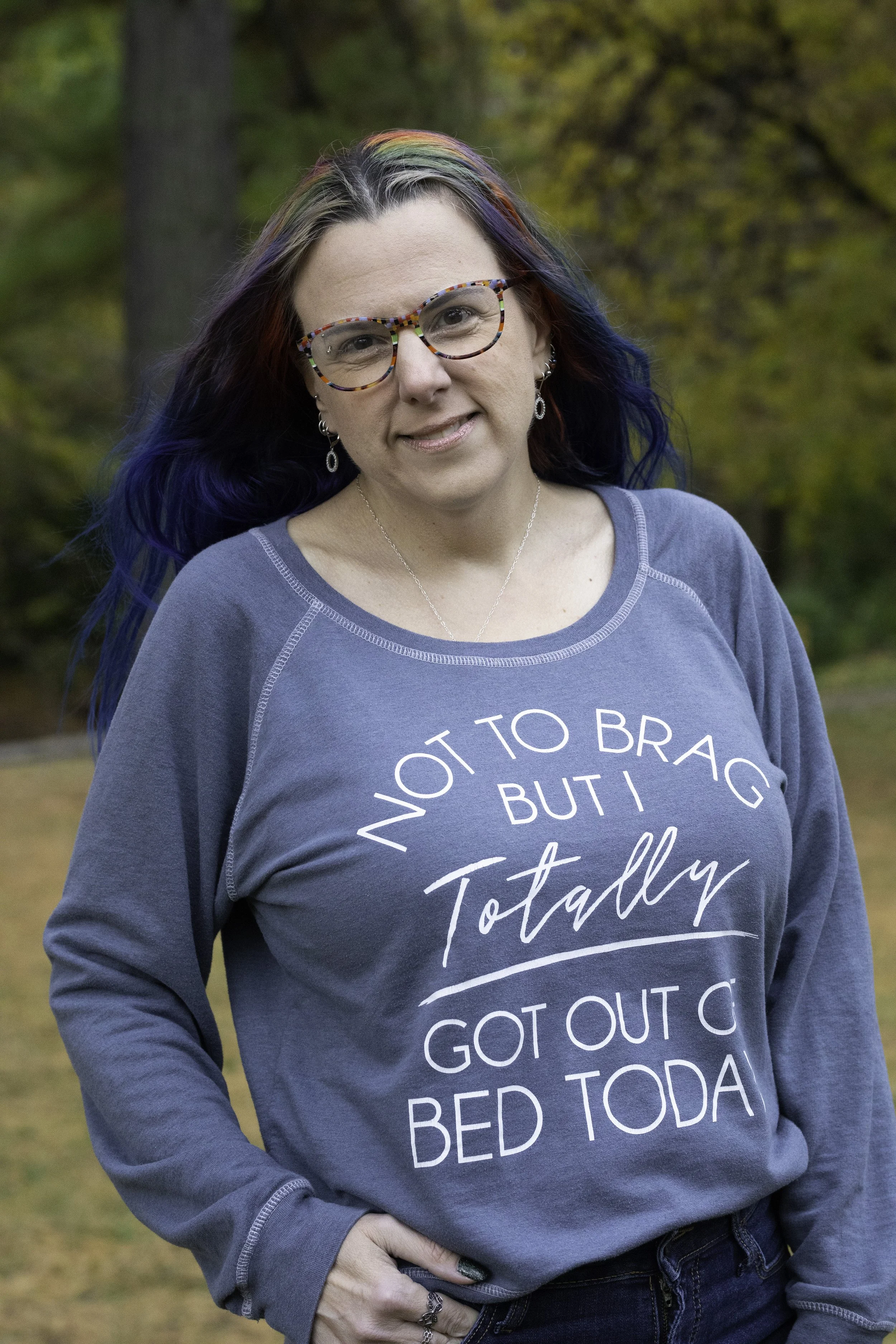 A woman with rainbow-colored hair and colorful glasses standing outdoors in a forested area wearing a blue sweatshirt that reads 'Not to brag but totally got out of bed today'.