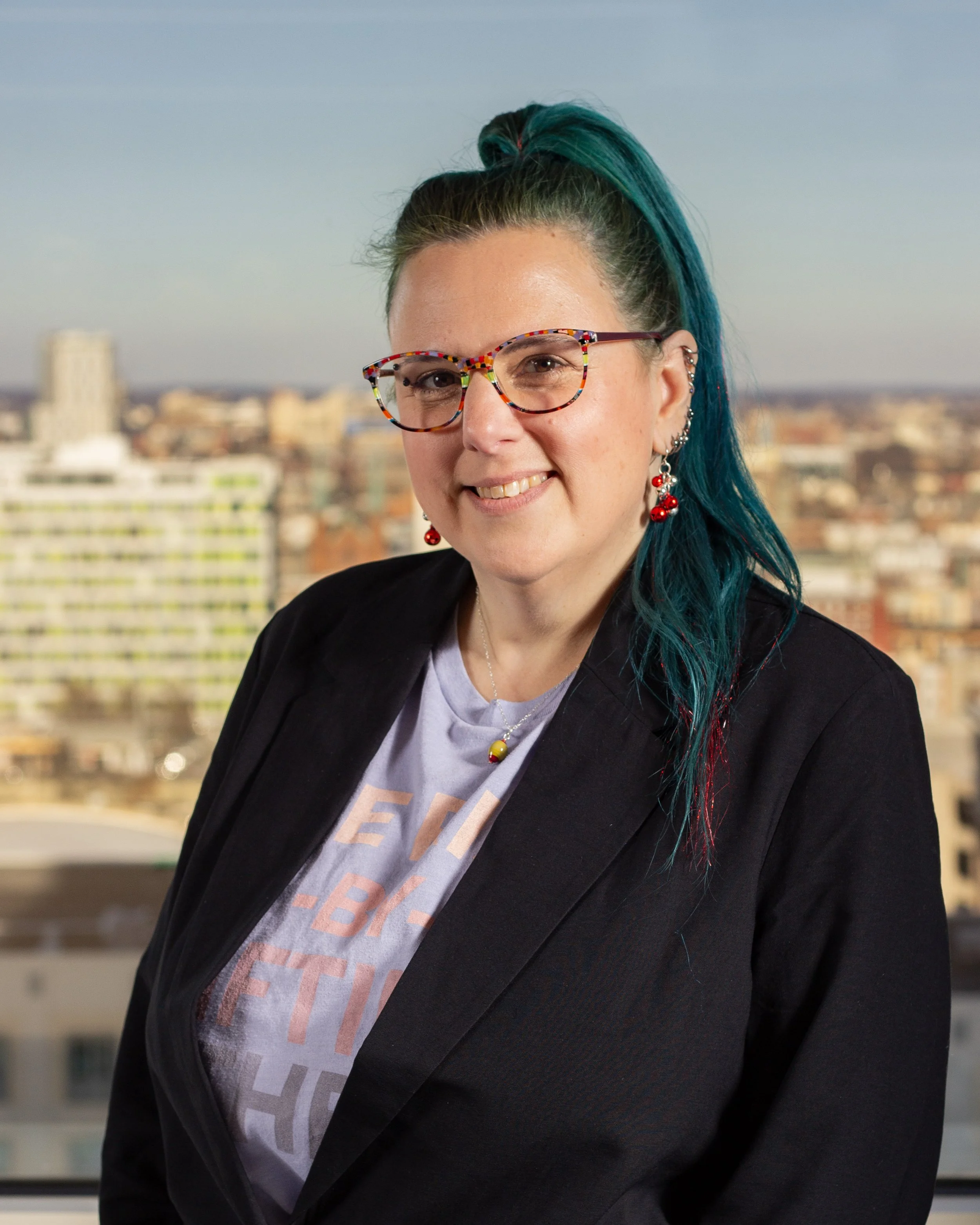 A woman with teal hair and colorful glasses smiling, wearing a black blazer and a graphic t-shirt, standing outdoors with a cityscape in the background.