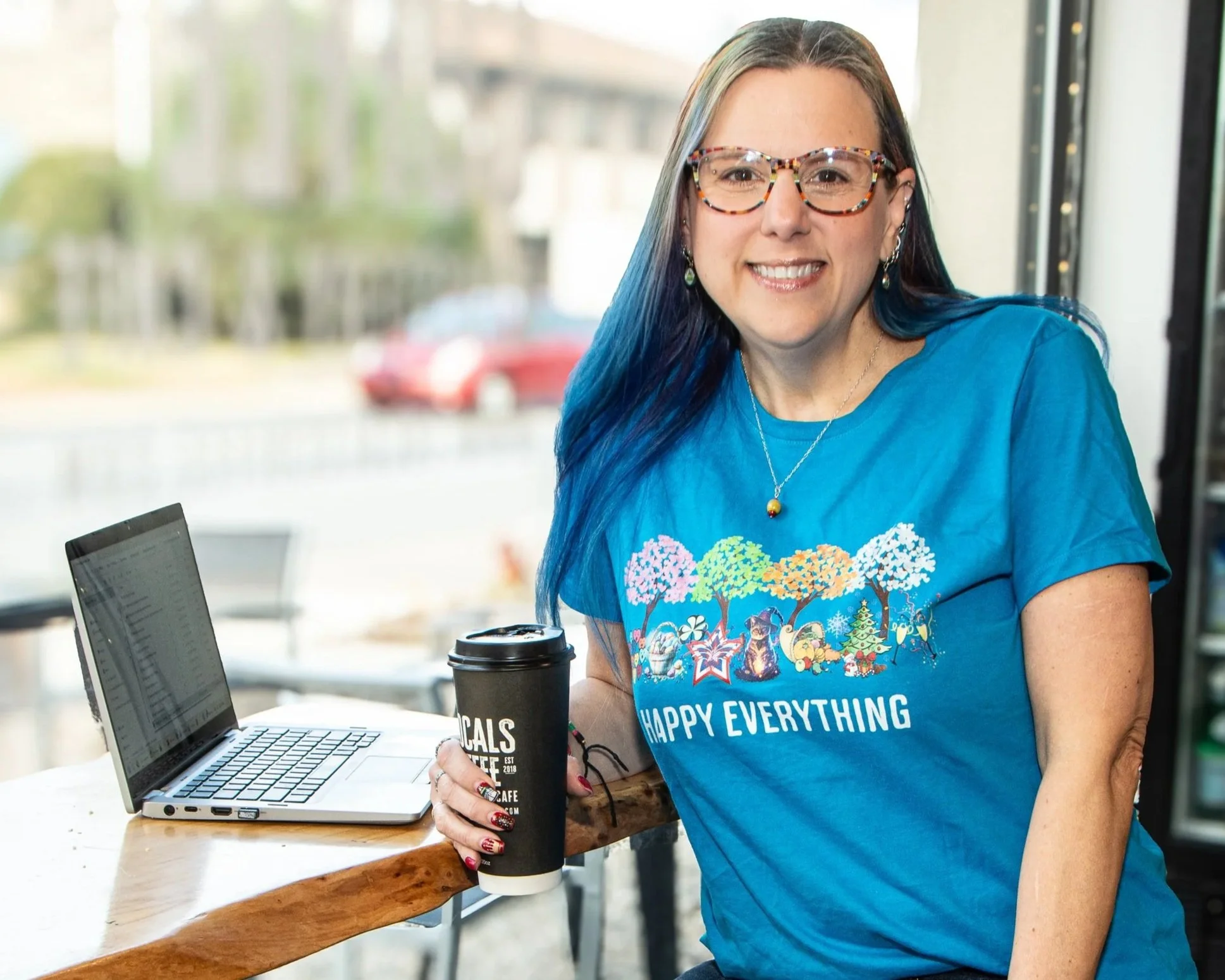 A woman with long colorful hair, glasses, and a bright smile sitting at a wooden table in a cafe, holding a to-go coffee cup, with a laptop open in front of her.