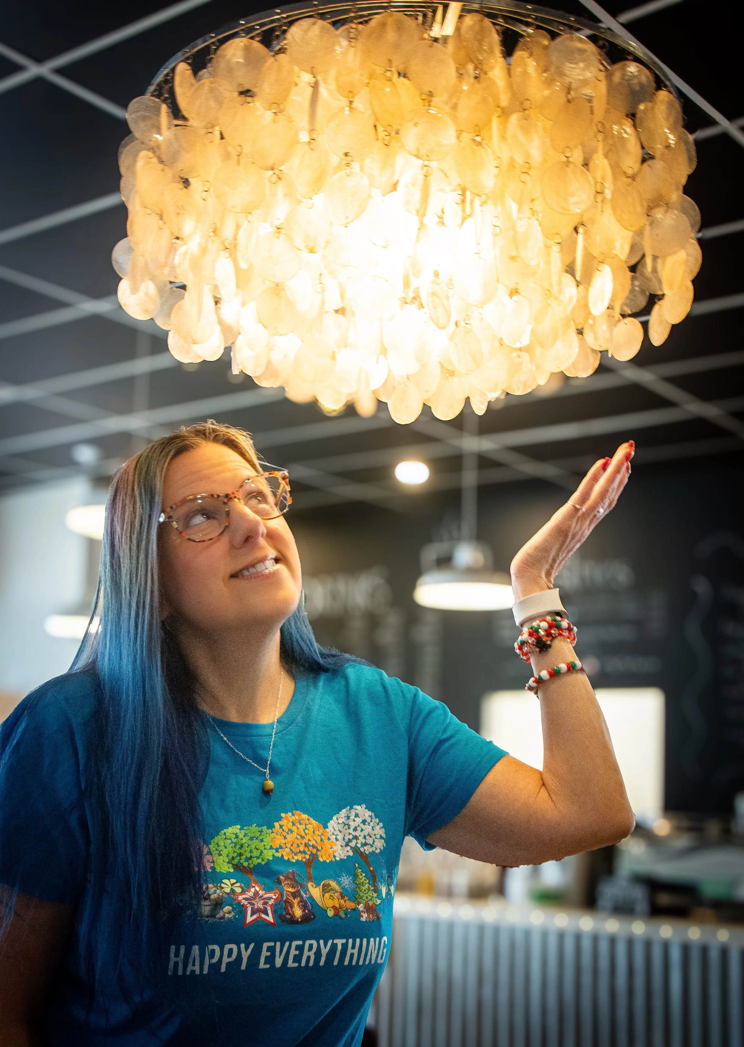 A woman with glasses and blue hair smiling and looking up at a chandelier made of shell-like discs.
