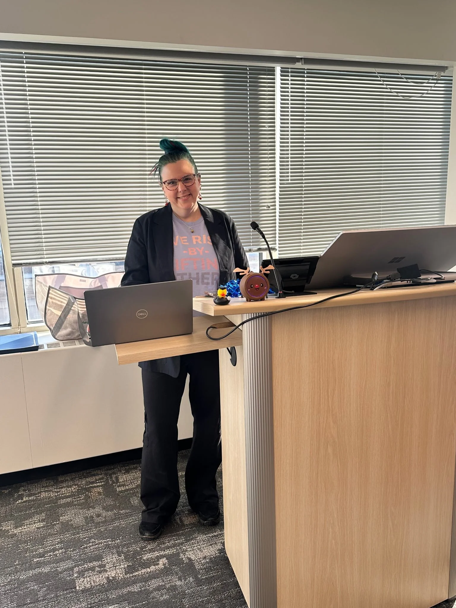 A woman standing at a desk with a Dell laptop, two additional monitors, colorful decorations, and a microphone, in a room with large windows and blinds.