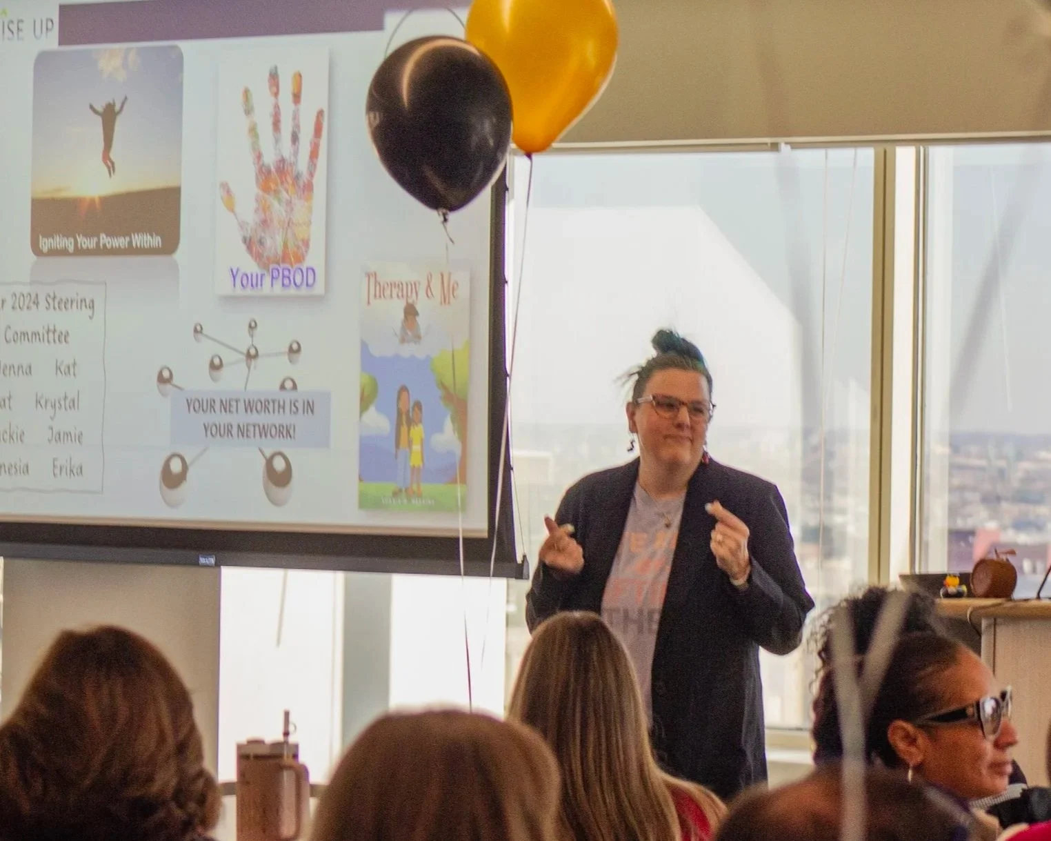 A woman giving a presentation in a conference room with large windows, with a slide showing images of a person jumping, a hand with colorful artwork, and children playing, along with balloons in the room.