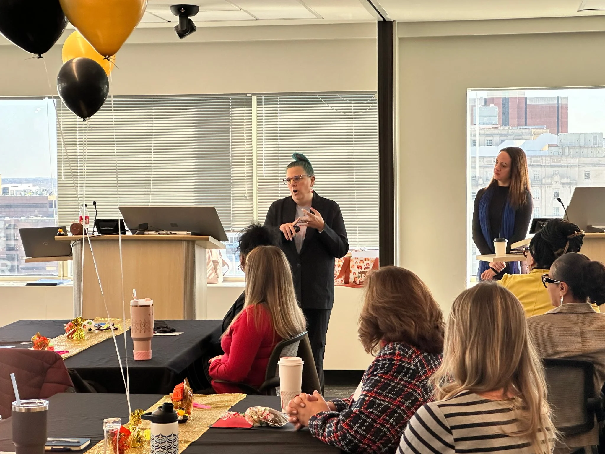 Women giving a presentation to a group in a conference room decorated with balloons and holiday-themed items.