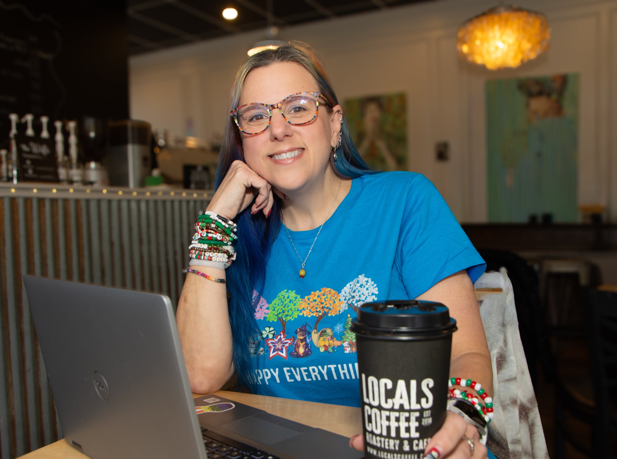 Woman sitting at a table in a cafe with a laptop, holding a coffee cup with "Locals Coffee" on it, wearing colorful bracelets, glasses, and a blue t-shirt, smiling at the camera.