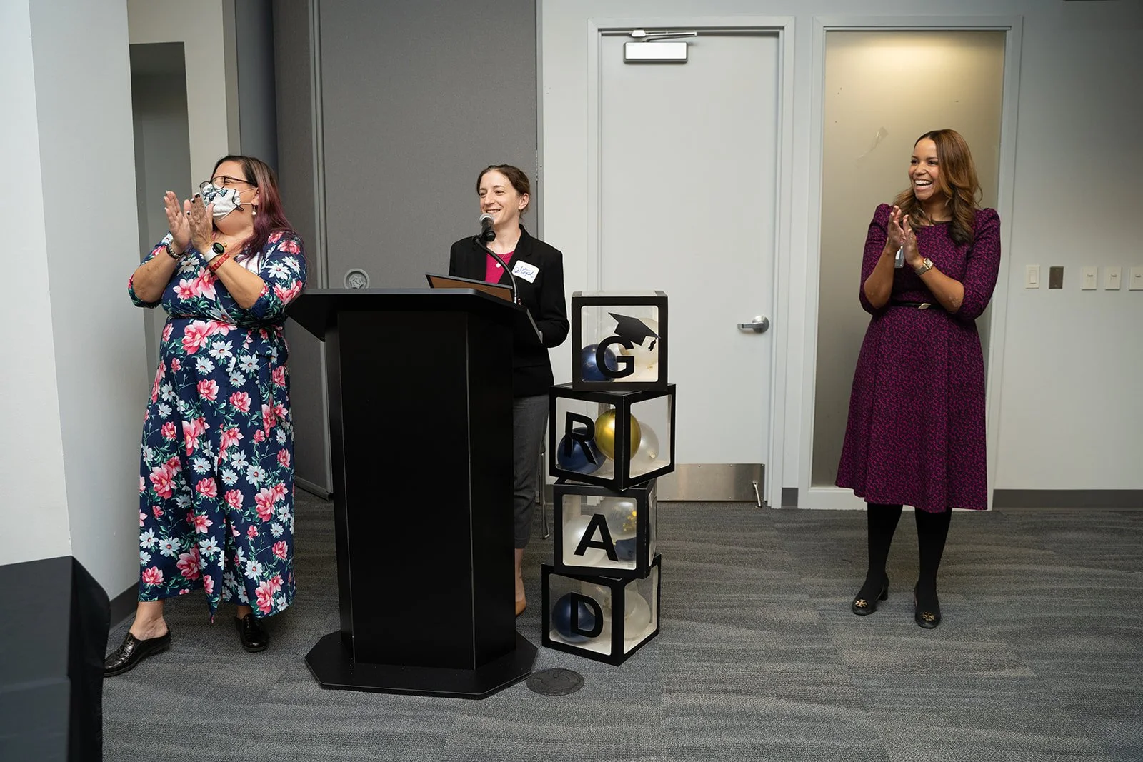 Three women standing in front of a grey wall at an indoor event. The woman on the left, wearing a floral dress and mask, is clapping. The woman in the middle, at a podium, is speaking into a microphone and smiling. The woman on the right, in a purple dress, is also clapping and smiling. There is a decorative display of black cube boxes with balloons and the word 'GRAD' arranged vertically.