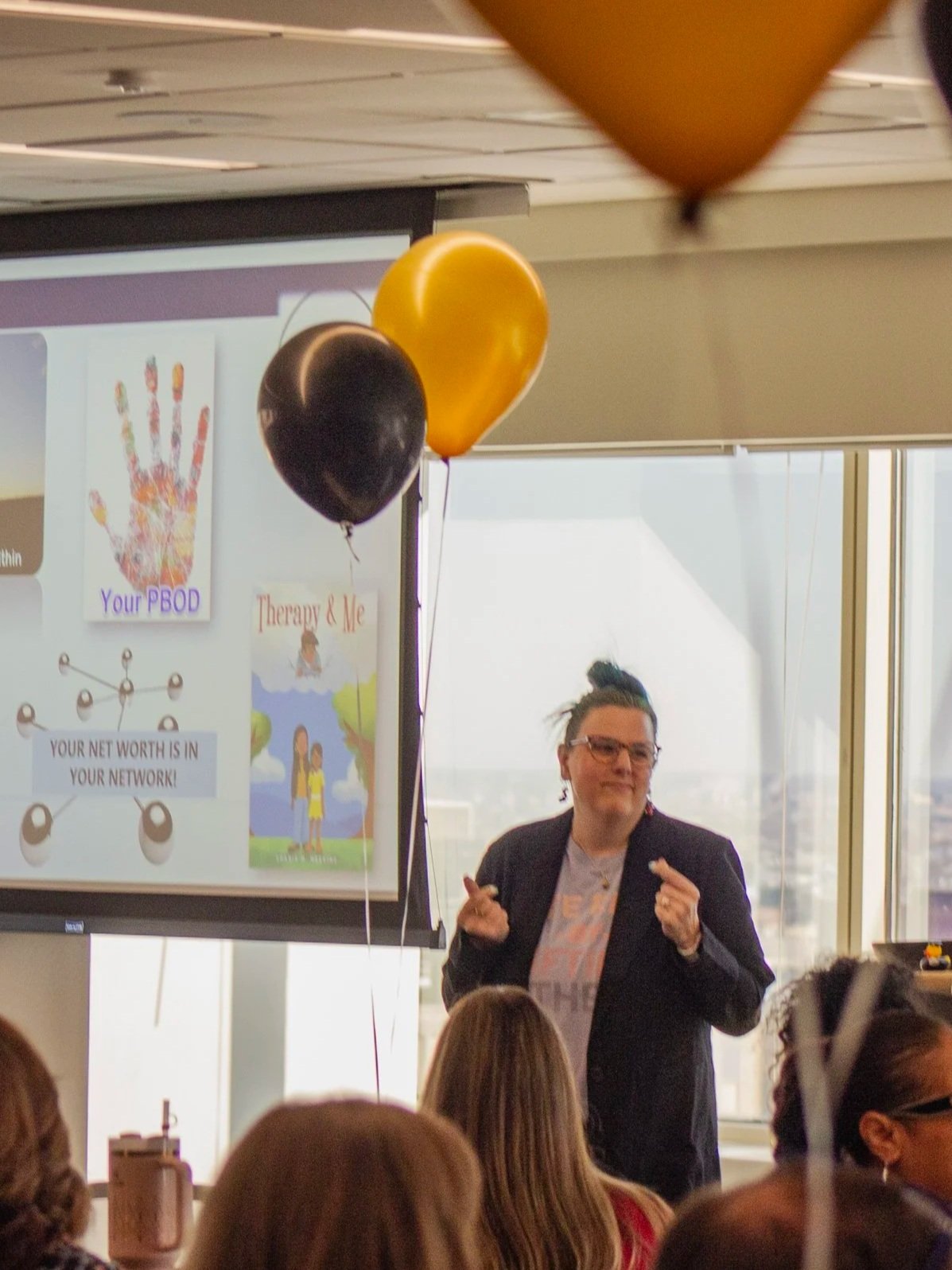 A woman giving a presentation in front of a screen with colorful posters, while audience members watch. There are black and yellow balloons tied to the screen.