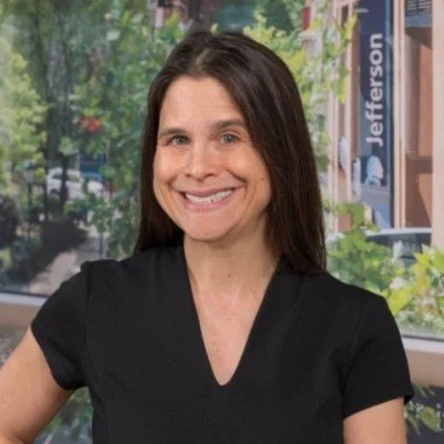 A woman with long dark hair smiling in front of a window with trees and buildings outside.
