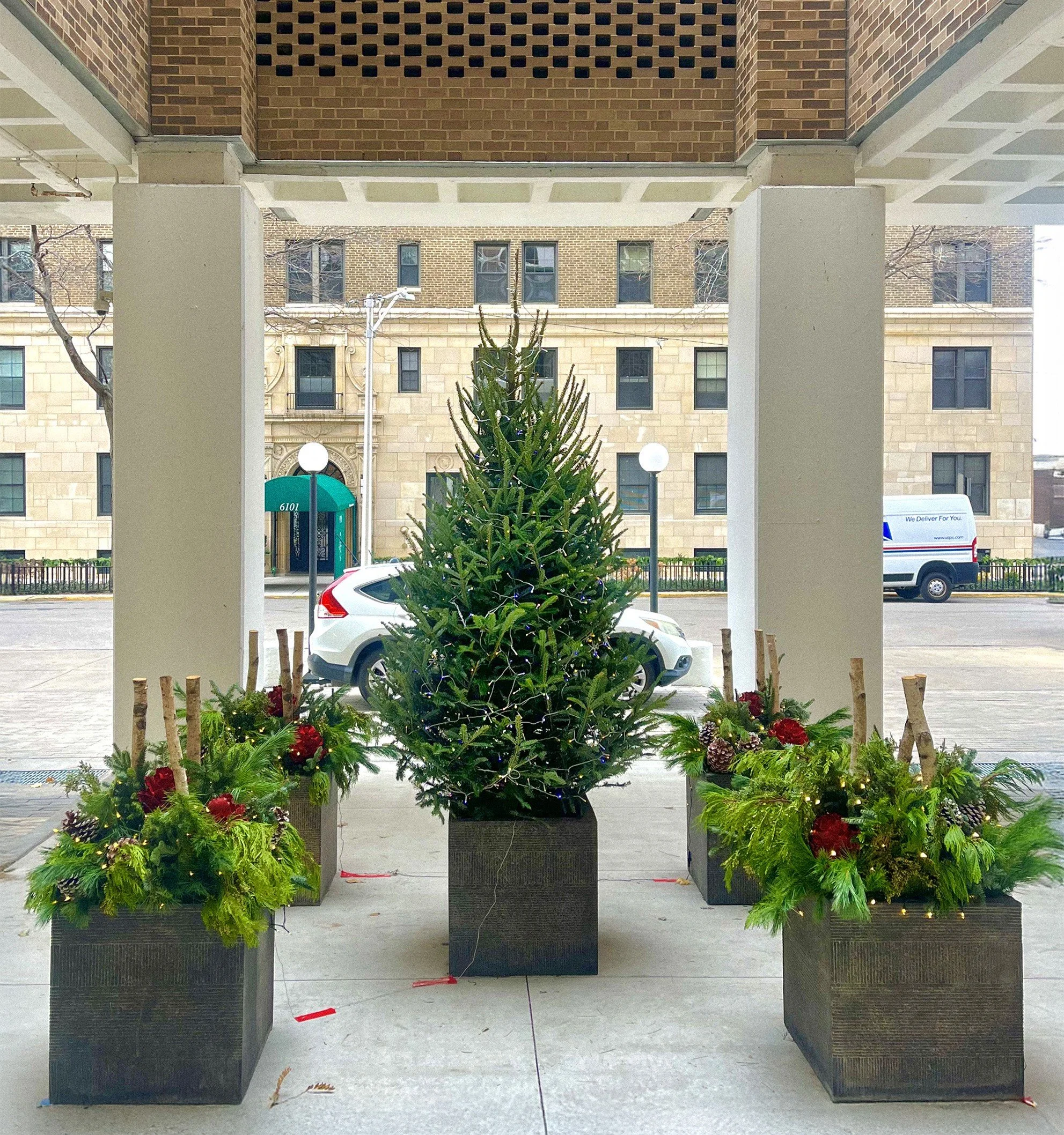 A decorated Christmas tree in a black planter with holiday arrangements on either side, located in an outdoor area with a building and parked cars in the background.