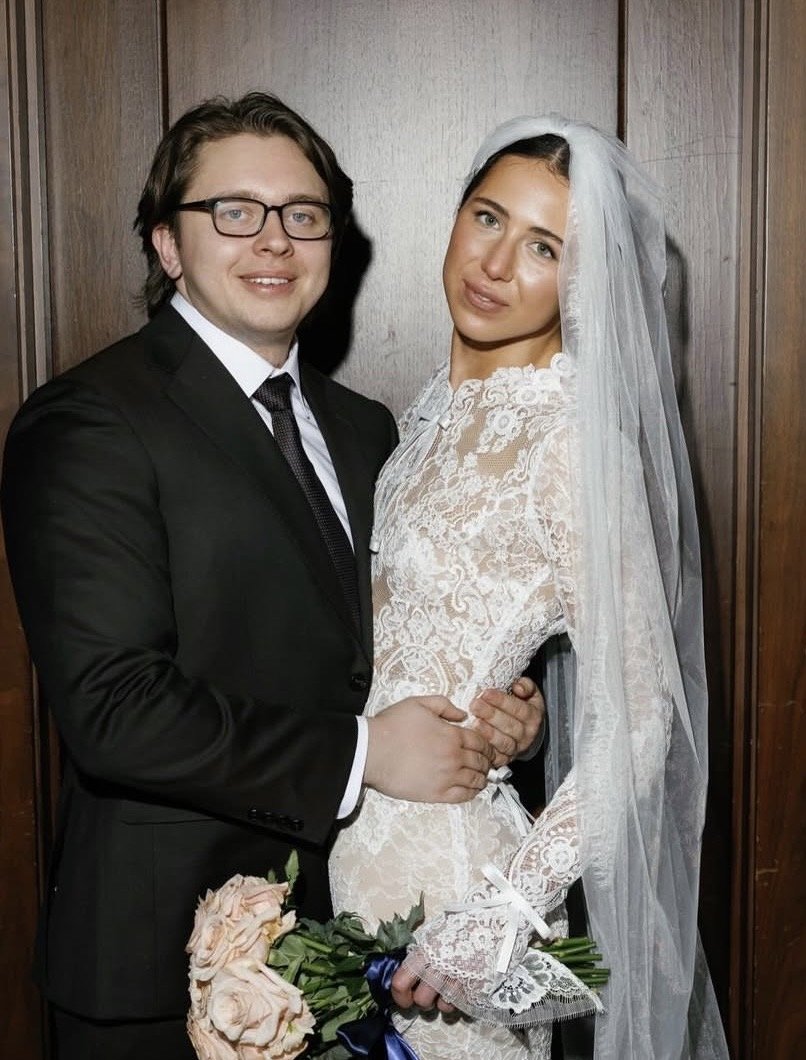 A newlywed couple in wedding attire posing on a city waterfront promenade with tall modern buildings in the background.
