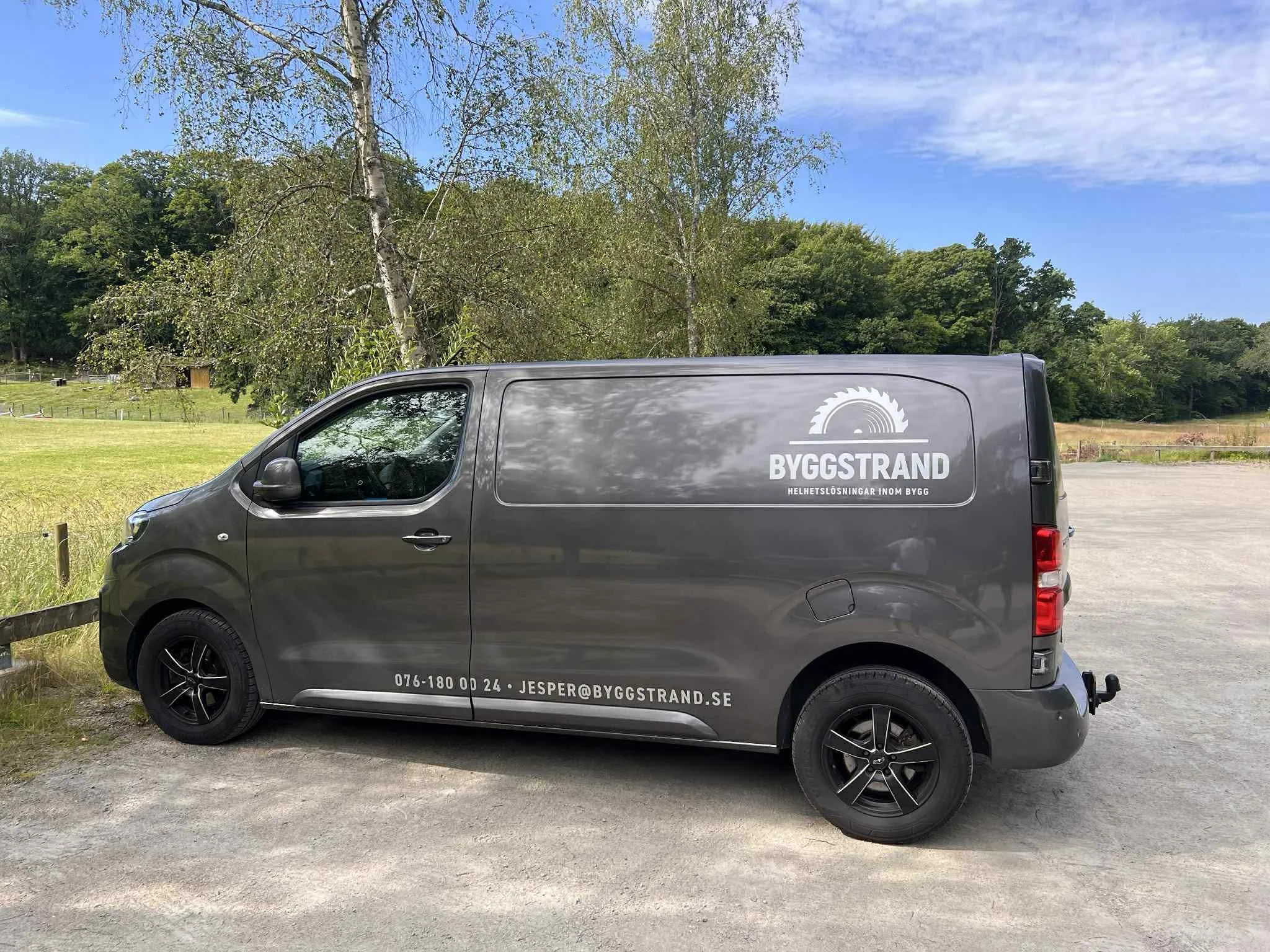 Grå företagsskåpbil parkerad på en grusplan med träd och ett gräsbevuxet fält i bakgrunden, med en logotyp och kontaktuppgifter.