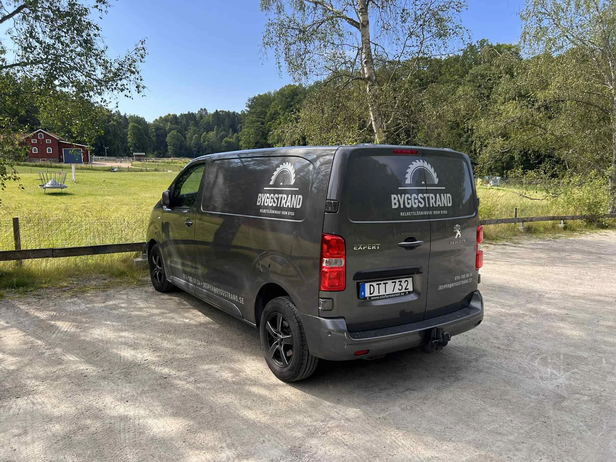En mörkgrå Peugeot Expert skåpbil med märkningen 'BYGGSTRAND' parkerad på en grusplan nära en gräsbevuxen åker och träd.