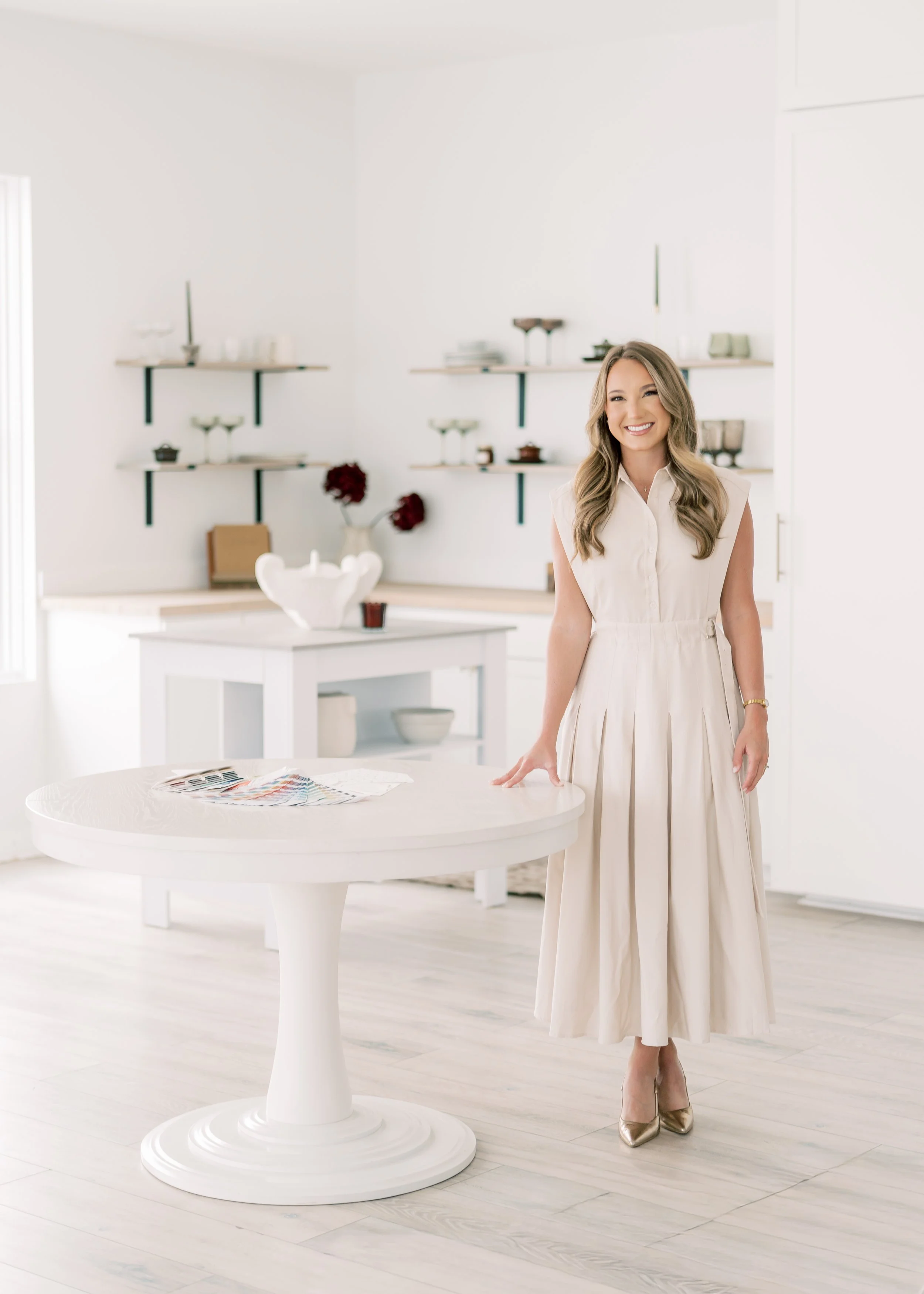 A woman in a cream-colored dress smiling and standing in a bright, minimalist kitchen with white walls, a round white table, and floating shelves with dishware and decorations.
