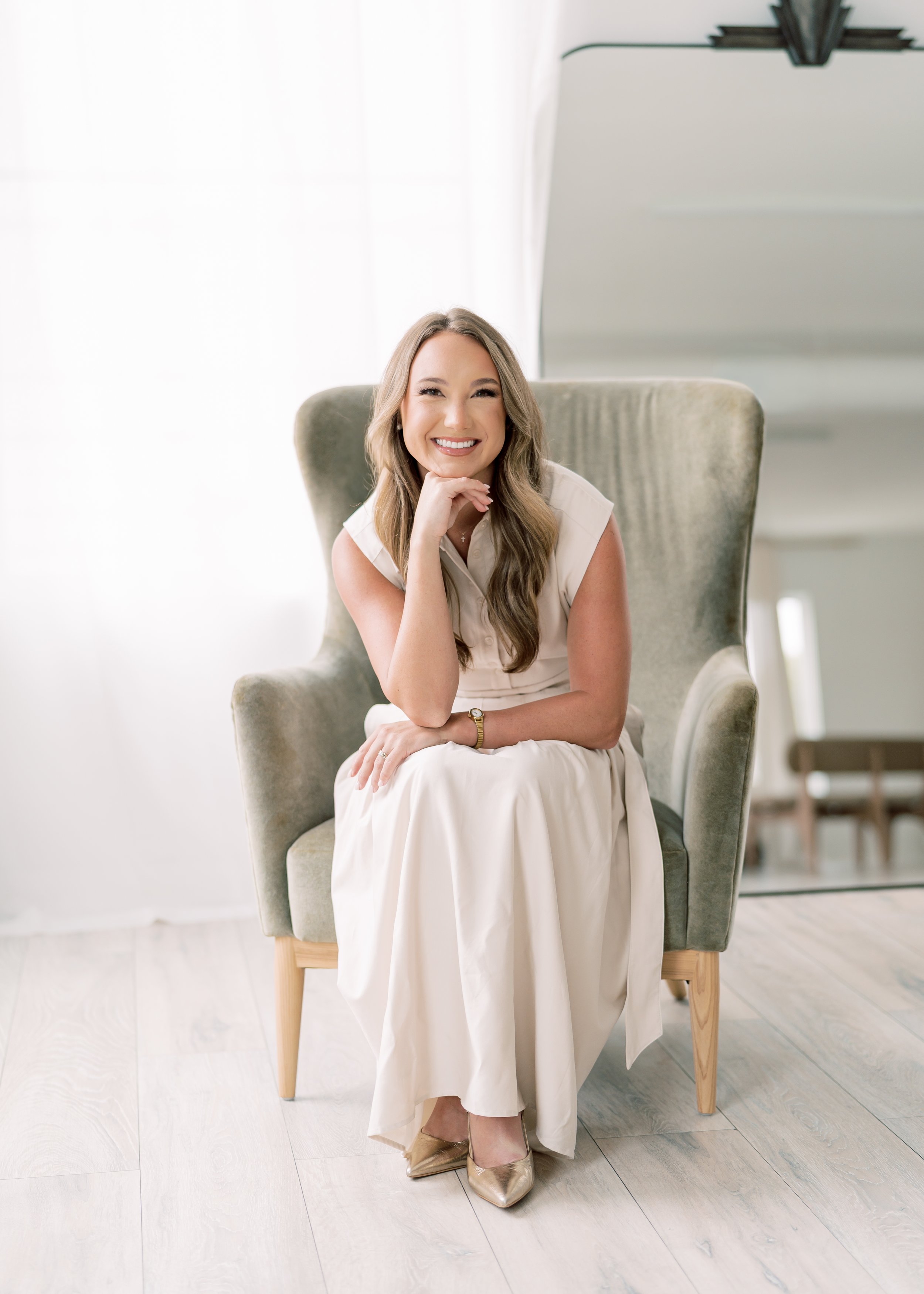 Smiling woman with long wavy hair sitting on a green velvet armchair in a bright room with white curtains and wooden flooring.