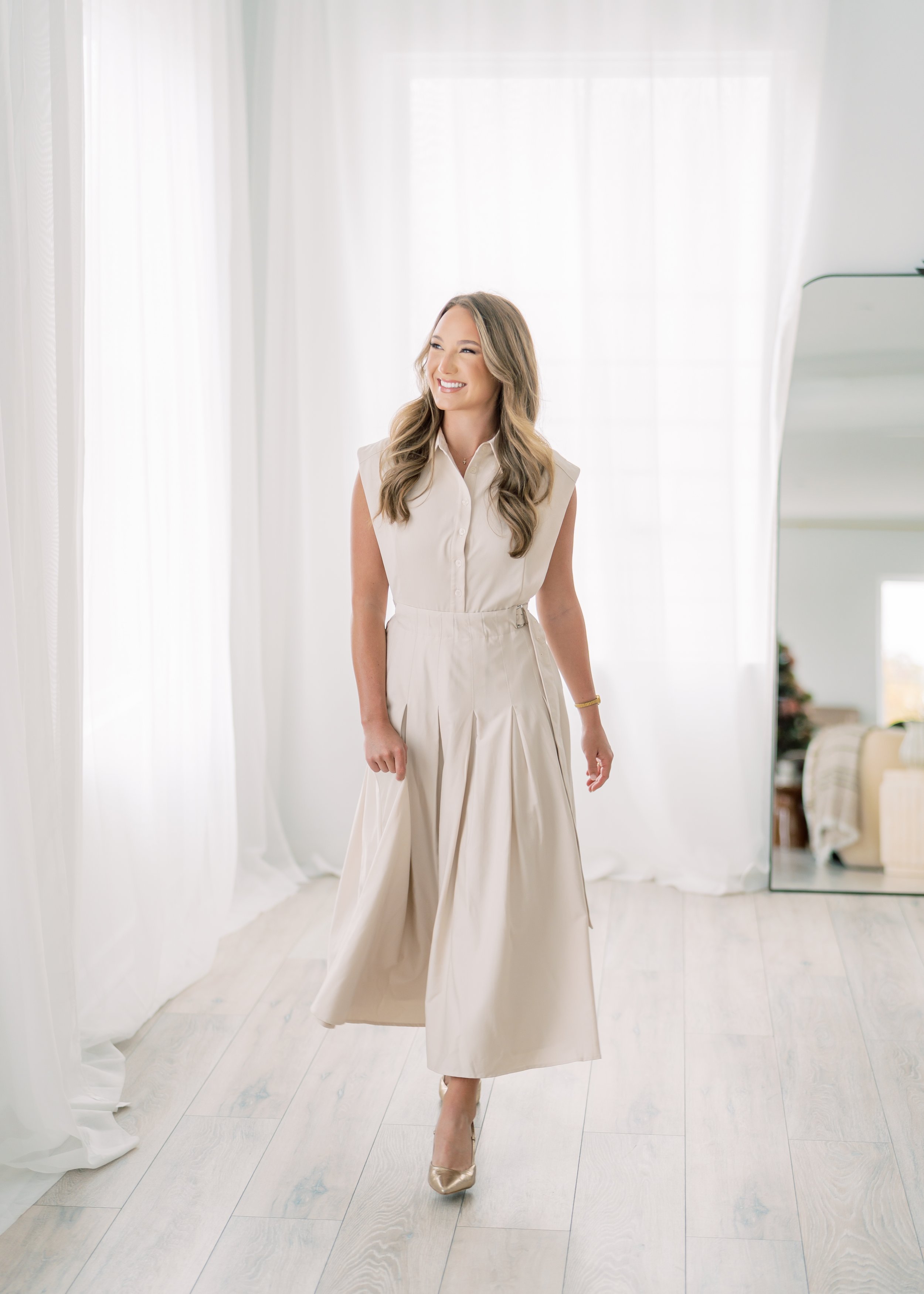 A woman in a cream-colored dress standing on a white wooden floor near sheer white curtains and a tall mirror, smiling.