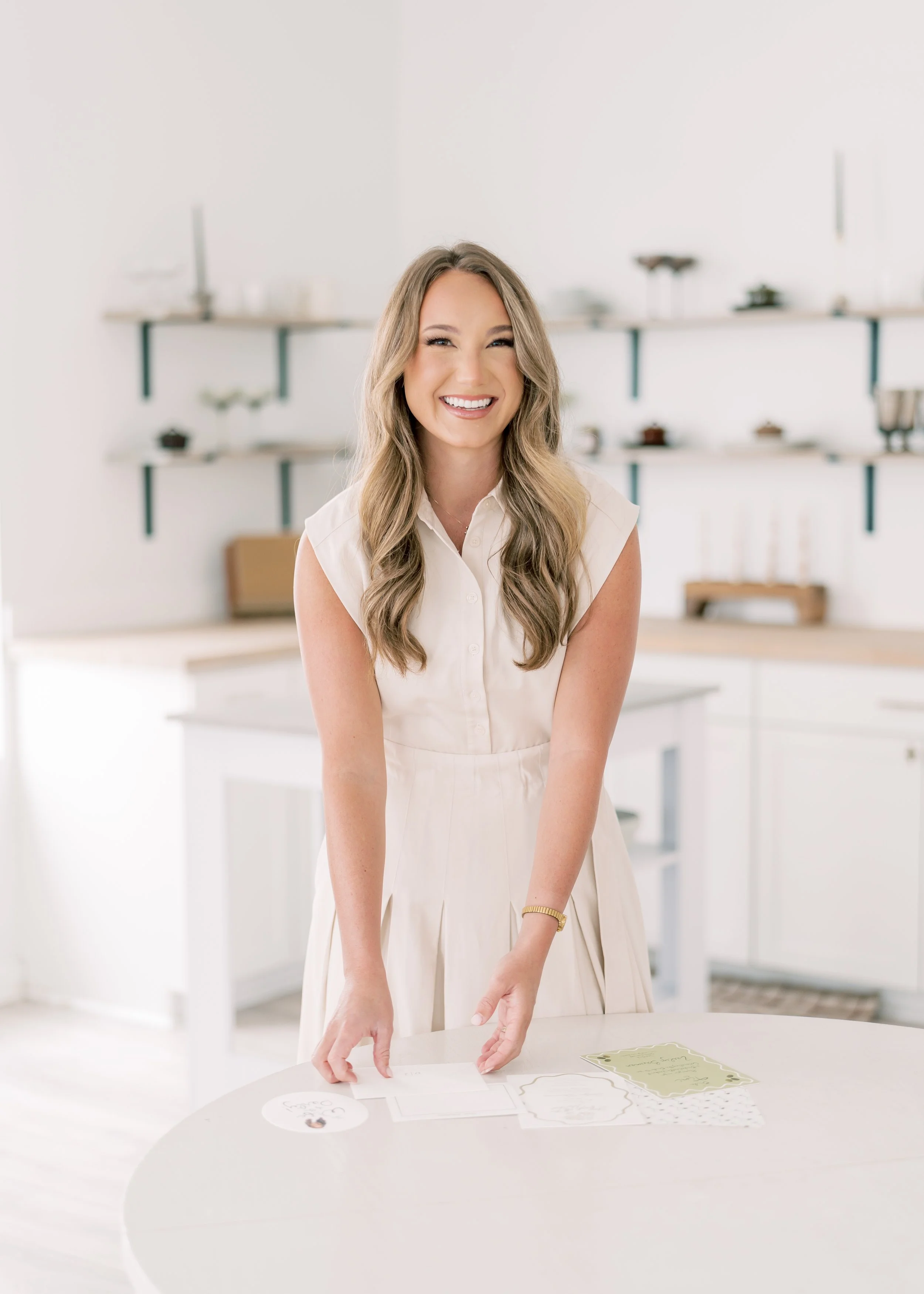 A woman with long wavy hair, wearing a cream-colored sleeveless dress, smiles at the camera while standing in a bright, modern kitchen with white cabinets and open shelves in the background.