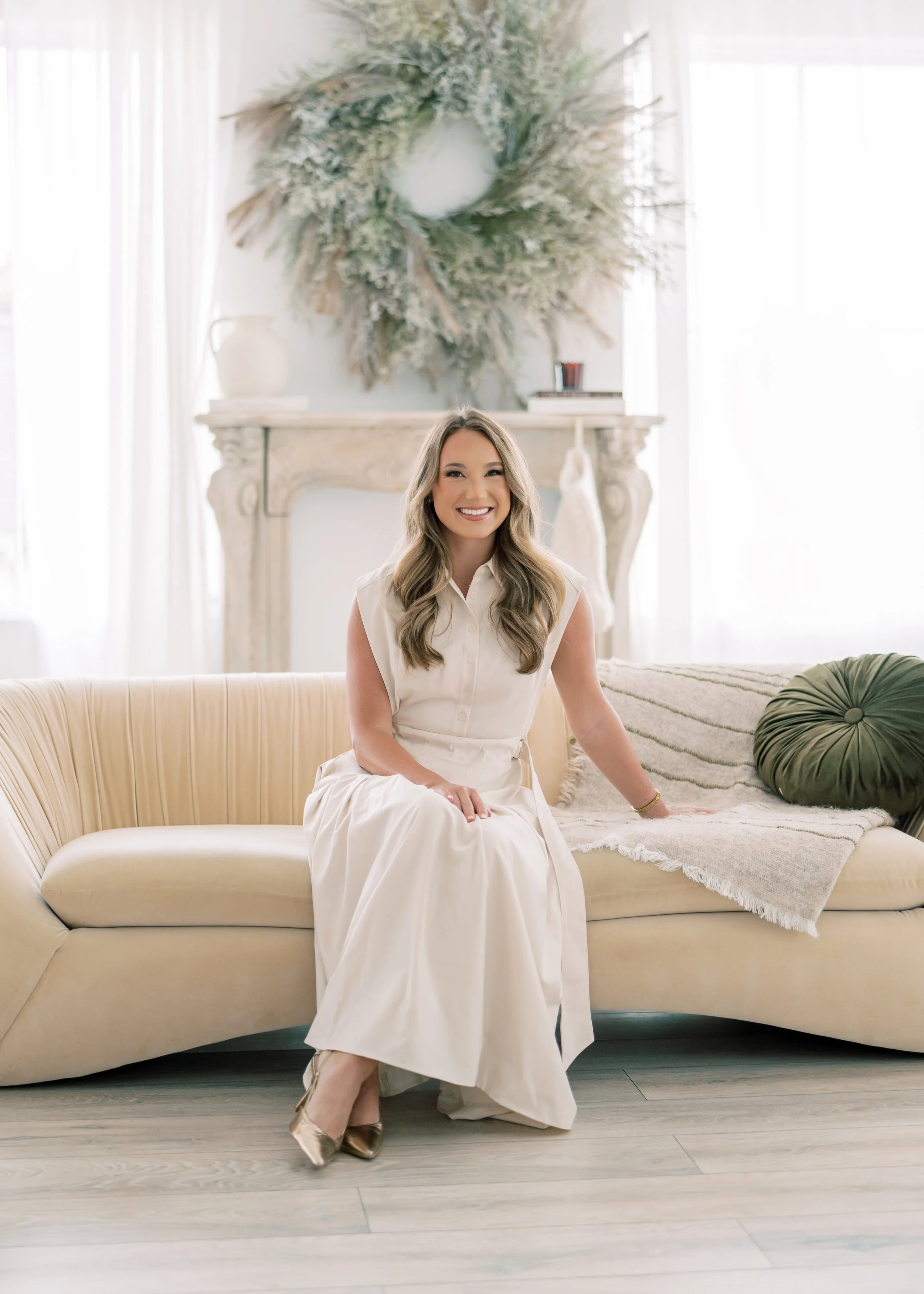 A woman in a cream-colored dress sitting on a beige sofa in a bright room with white curtains, a decorative fireplace, and a large wreath hanging above it.