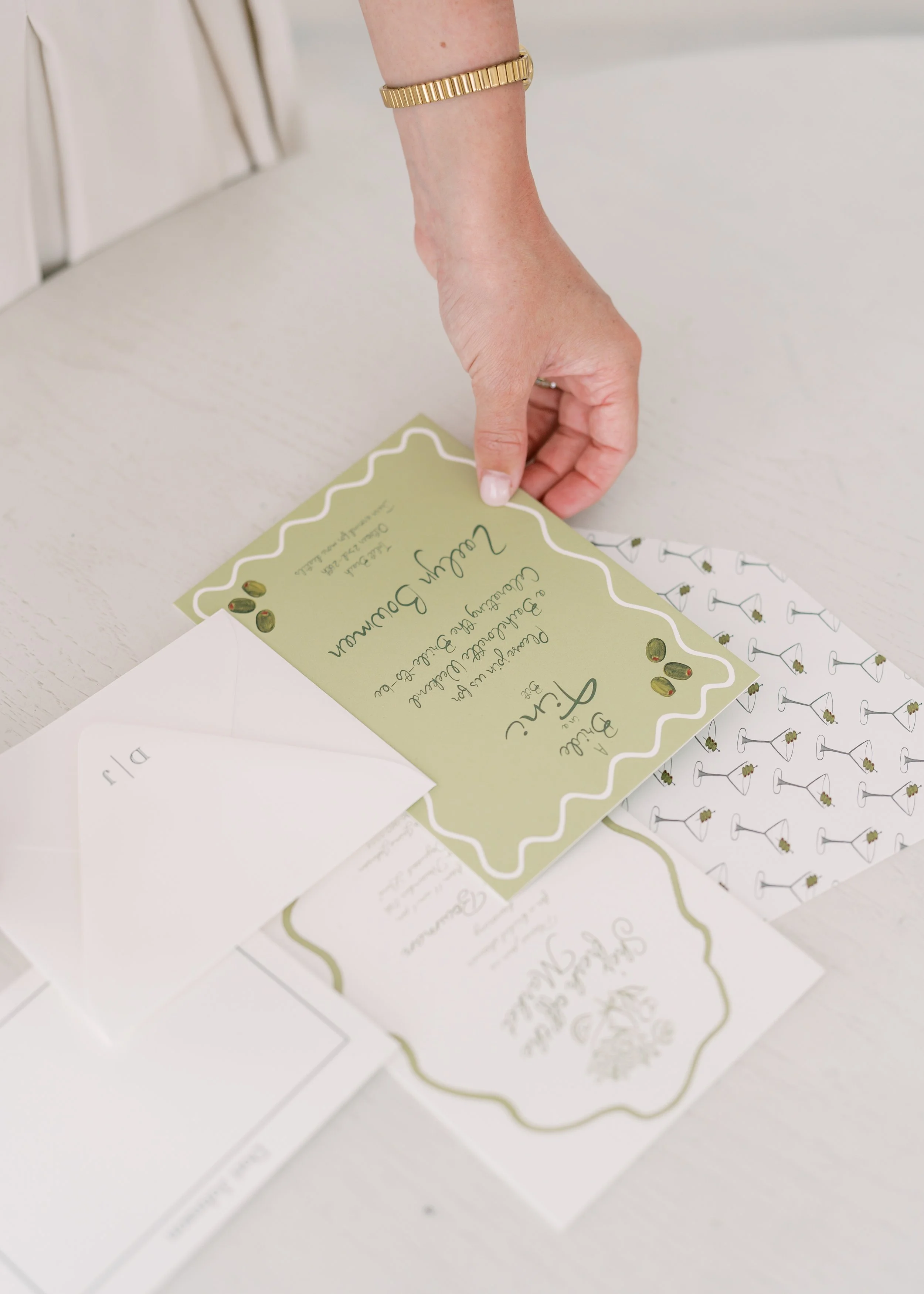 A woman with a gold bracelet places a green and white wedding invitation on a white table, with other invitations and envelopes nearby.