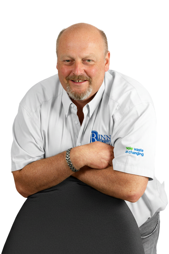 A smiling middle-aged man with a goatee and bald head, wearing a white button-up shirt with logos, leaning on a black surface with crossed arms, against a plainwhite background.