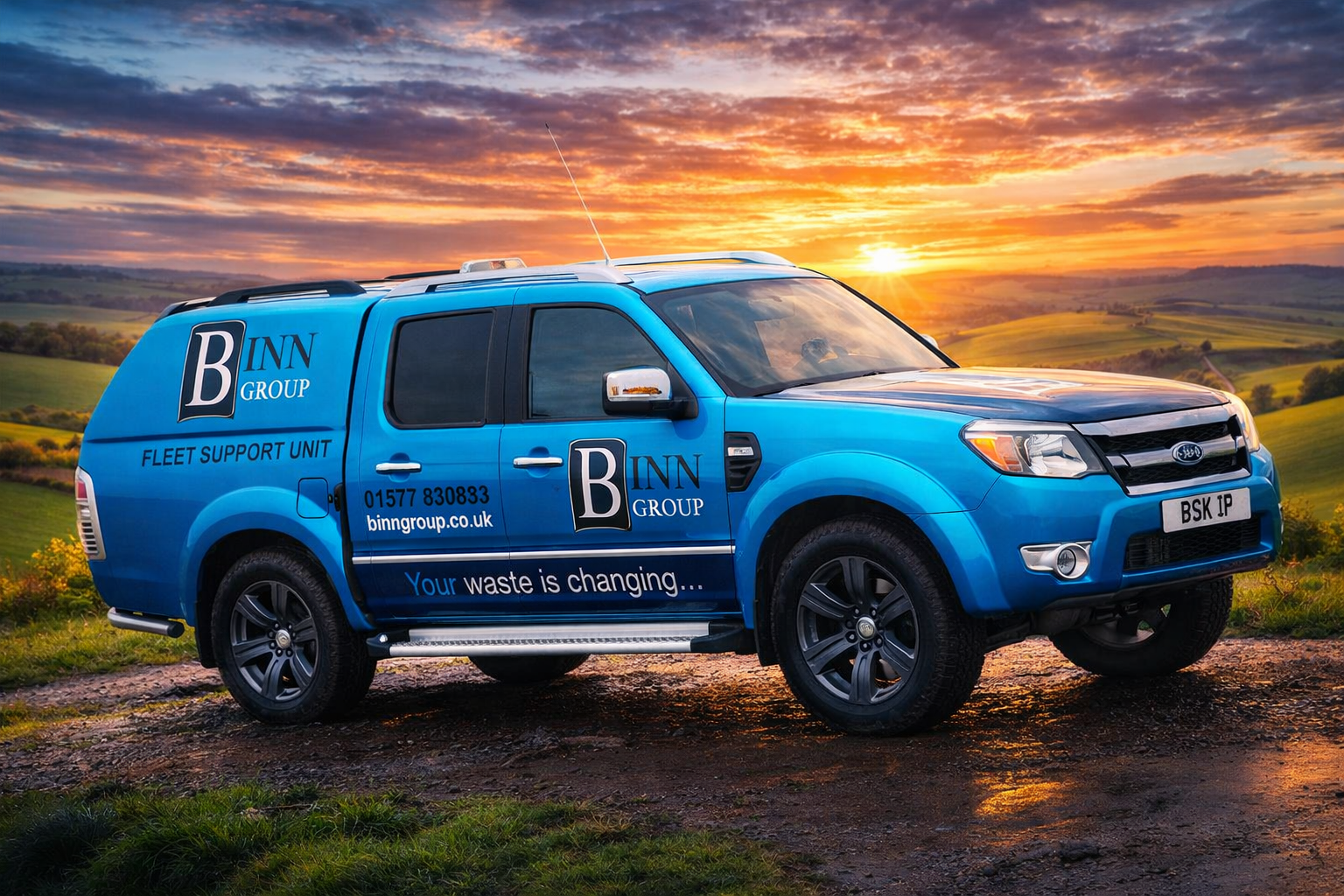 A blue fleet support unit vehicle with 'BINN GROUP' branding parked on a dirt road with a scenic sunset and rolling green hills in the background.