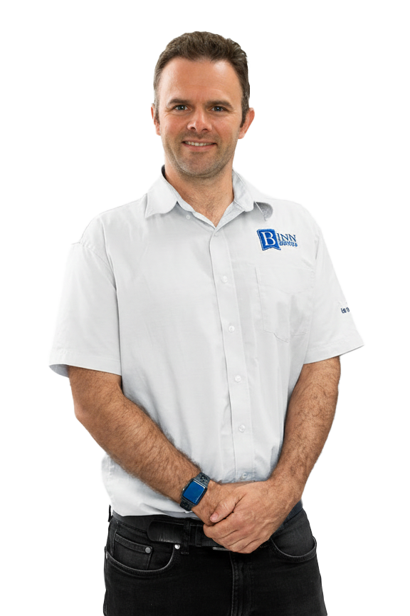 A man with short brown hair and light skin, smiling, wearing a white short-sleeved collared shirt with a logo, standing with his hands clasped in front of him against a white background.