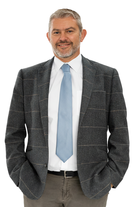 A middle-aged man with gray hair and a beard, dressed in a dark gray plaid blazer, white shirt, and light blue tie, smiling and posing with hands in pockets.