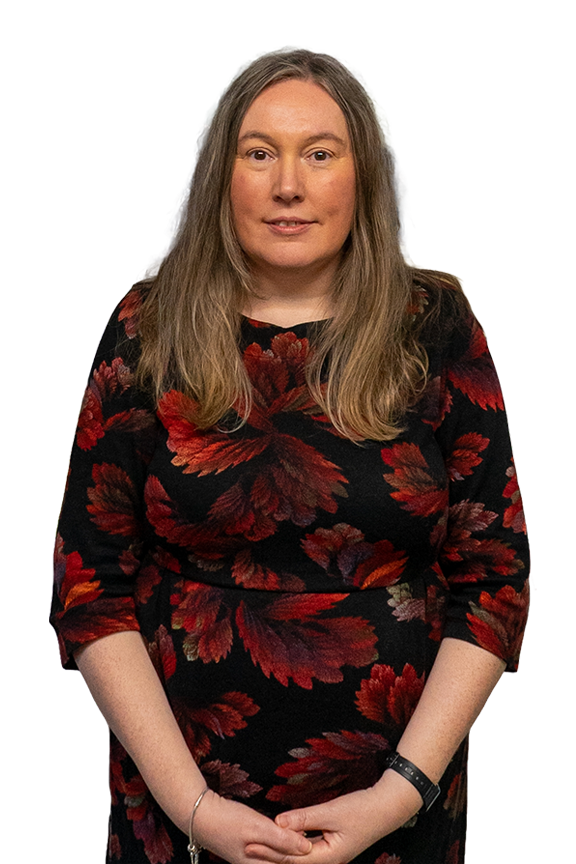 A woman with long, wavy, light brown hair, wearing a black dress with red and orange floral patterns, stands against a plain background.
