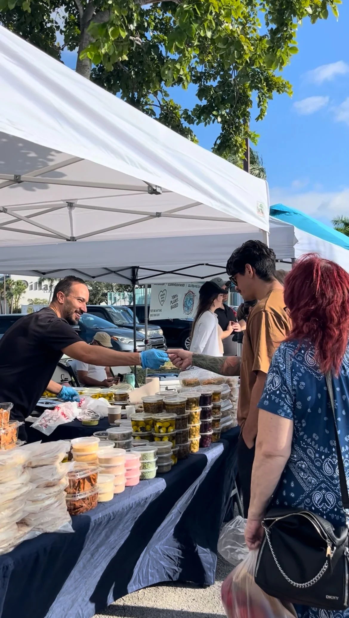 Vendor at outdoor market stall handing food sample to customer, with stacked containers of dips, spreads, and snacks on table, under white canopy with people and vehicles in background.