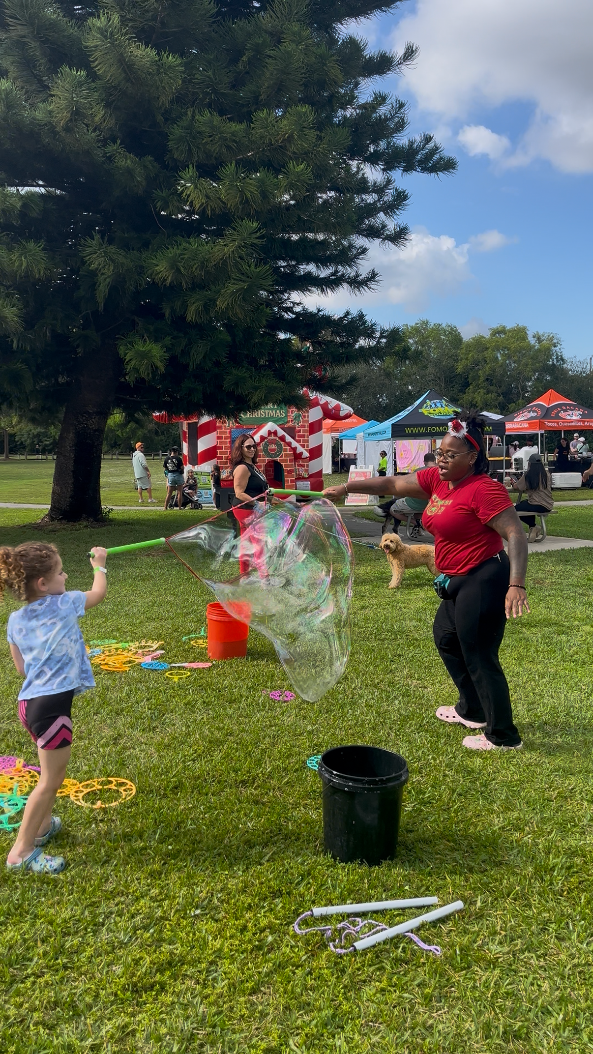 A young girl is blowing a large bubble with a bubble wand while an adult woman helps with the bubble wand, at a park during a festive outdoor event with tents, people, and a large Christmas-themed inflatable in the background.