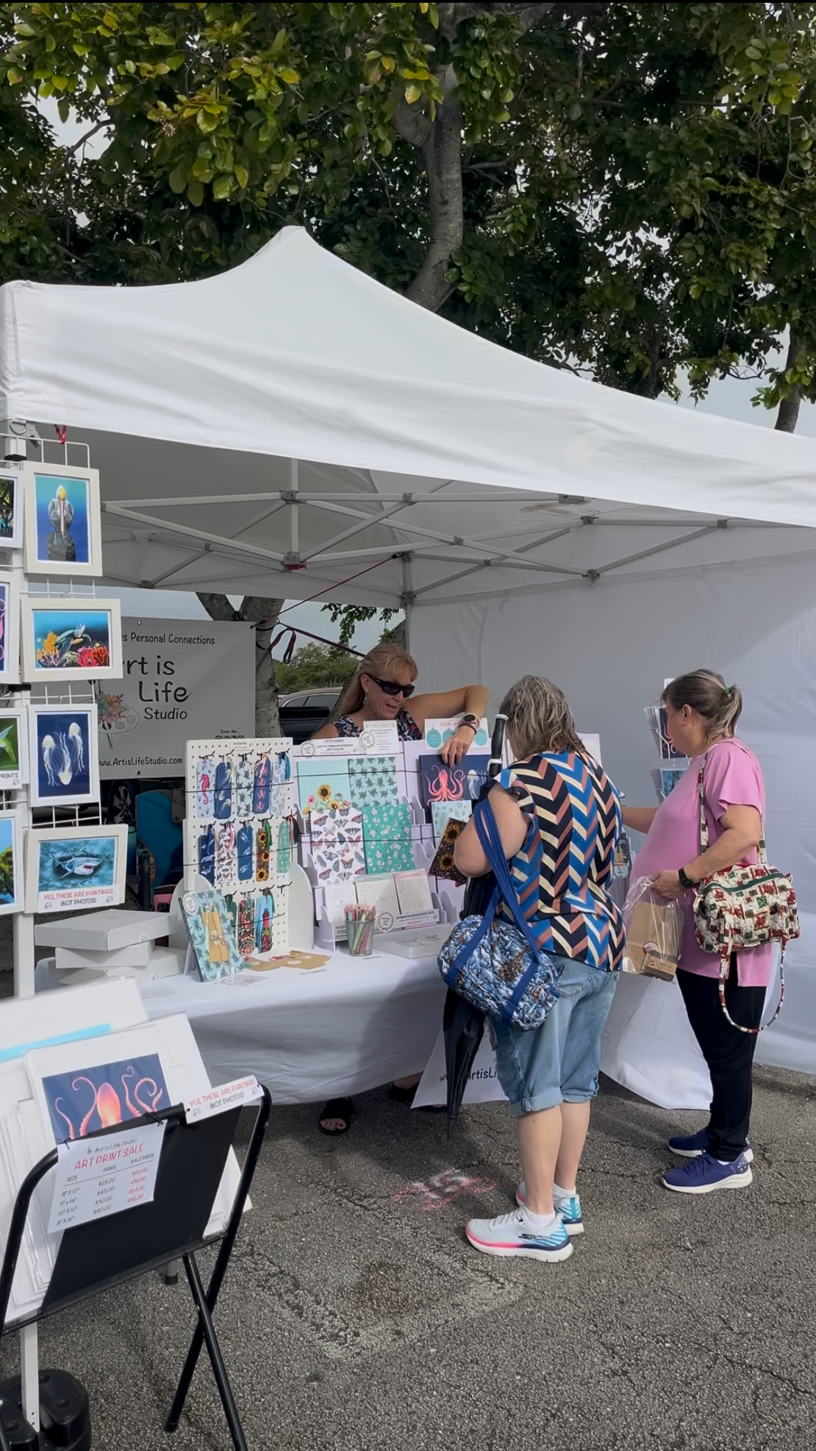Art vendors selling paintings and crafts under a white canopy at an outdoor market, with three women browsing and a sign for artwork and art print sale.