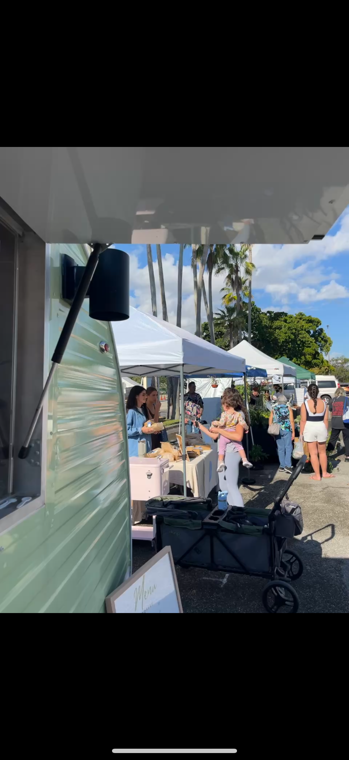Outdoor market scene with white tents, people shopping, and palm trees in sunny weather. A woman is carrying a child while browsing stalls.