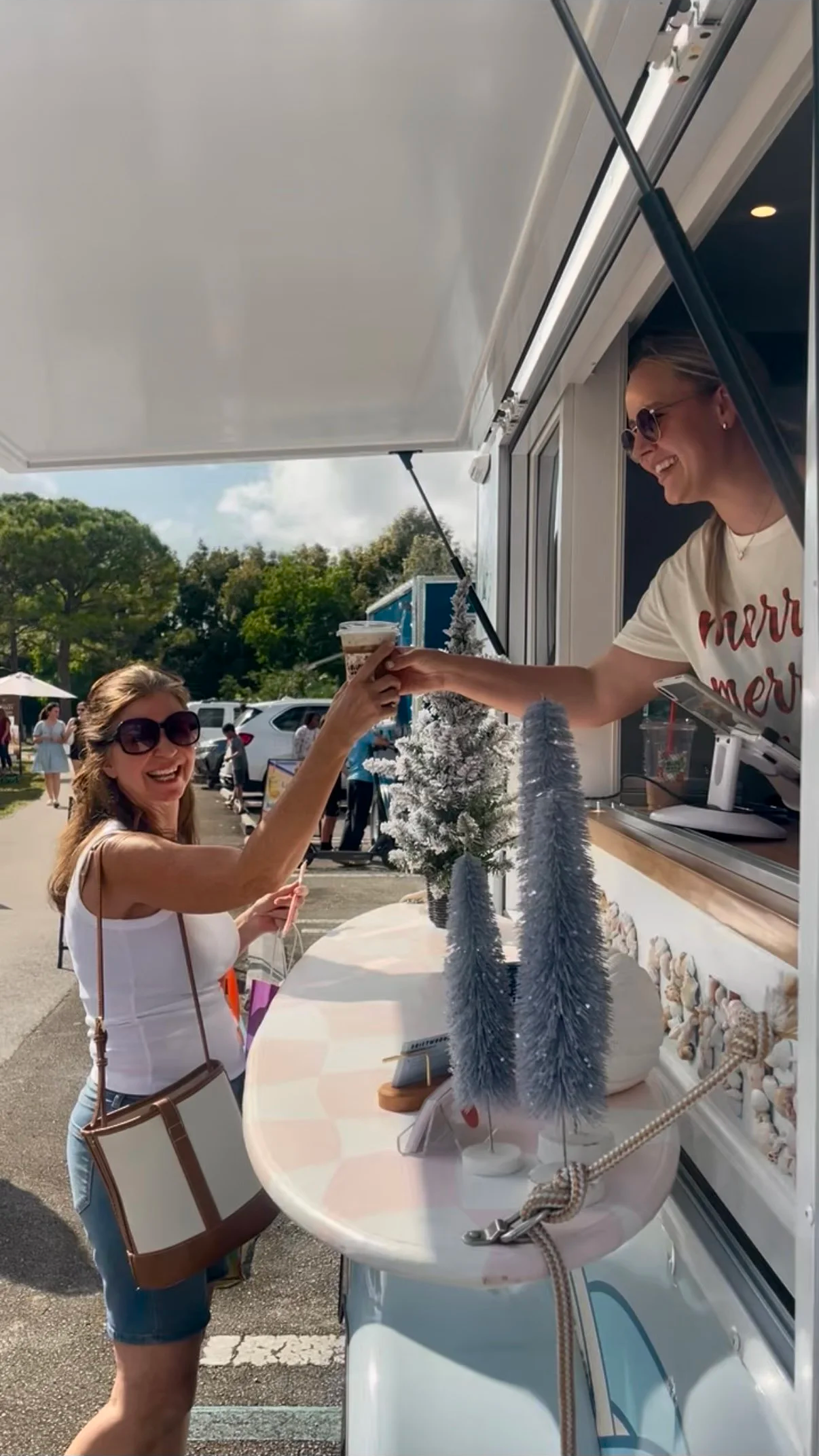 A woman in sunglasses is receiving a coffee from a smiling woman in a food truck at an outdoor event, with trees and parked cars in the background.