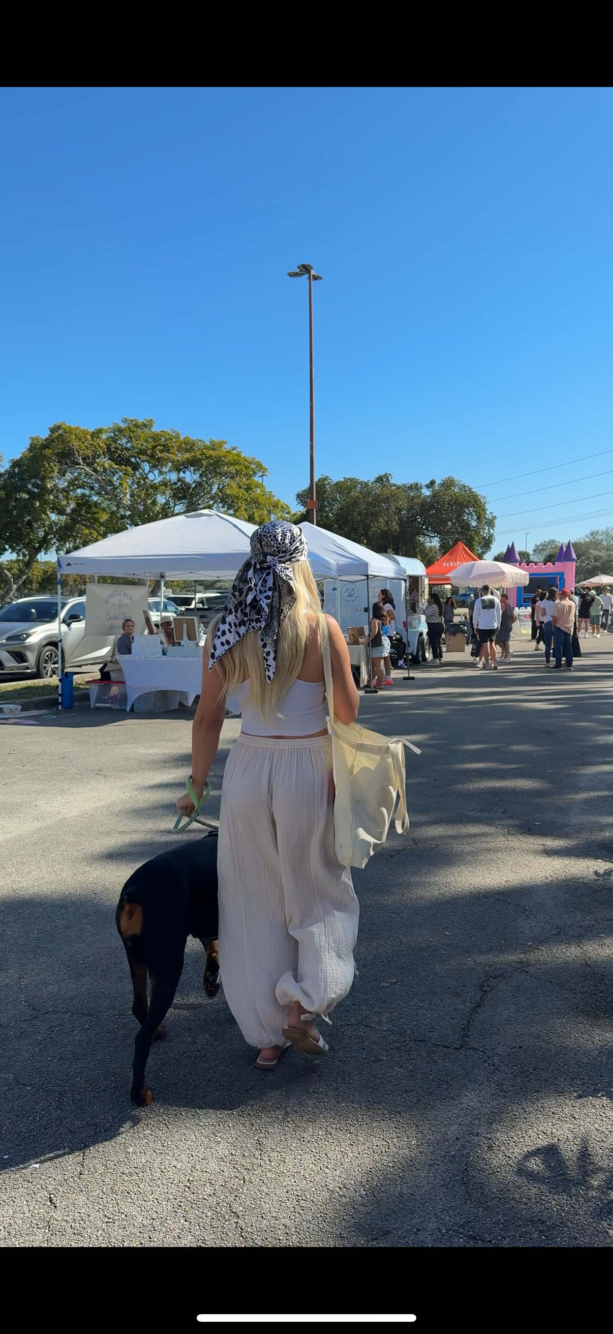 A woman with long blonde hair wearing a white top, beige pants, and a black-and-white patterned headscarf walks in an outdoor market, holding a beige bag and a leash attached to a black and brown dog. Behind her, market stalls with tents and people browsing are visible under a clear blue sky.