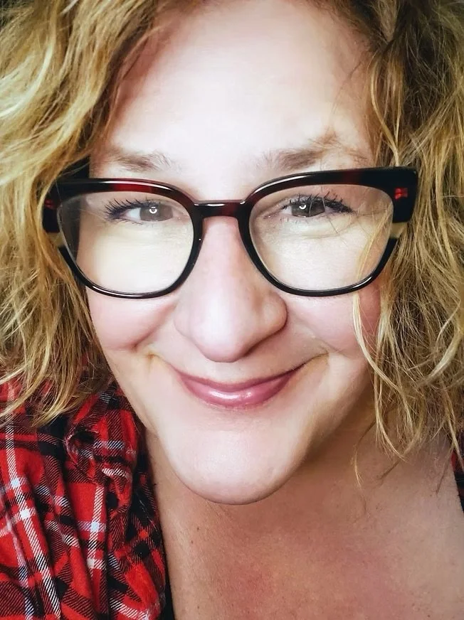 A close-up of a woman with curly blonde hair, wearing black glasses, smiling, and dressed in a red plaid shirt.