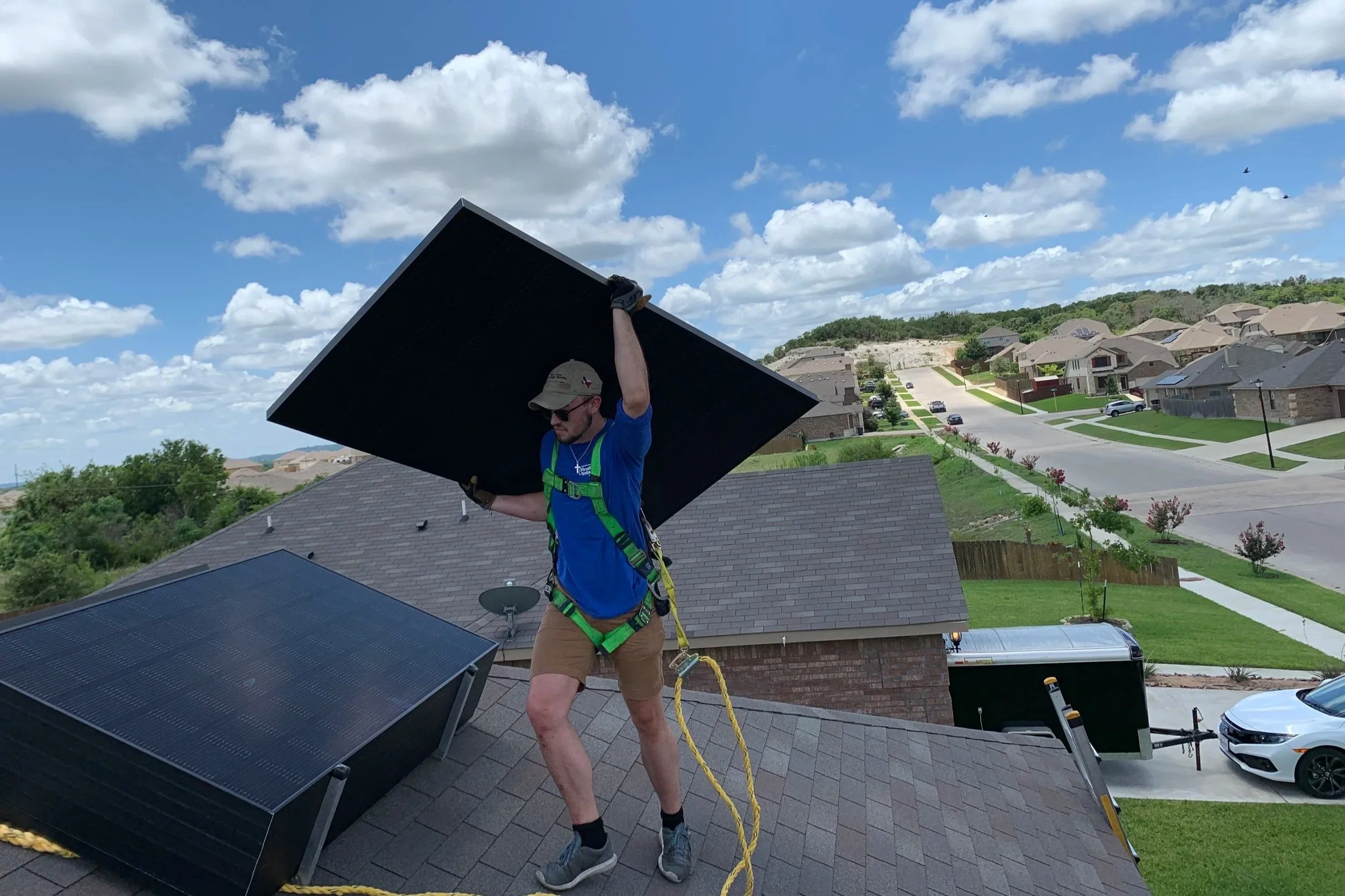 A man on a rooftop installing or adjusting a solar panel, wearing a harness and gloves, with a suburban neighborhood and partly cloudy sky in the background.