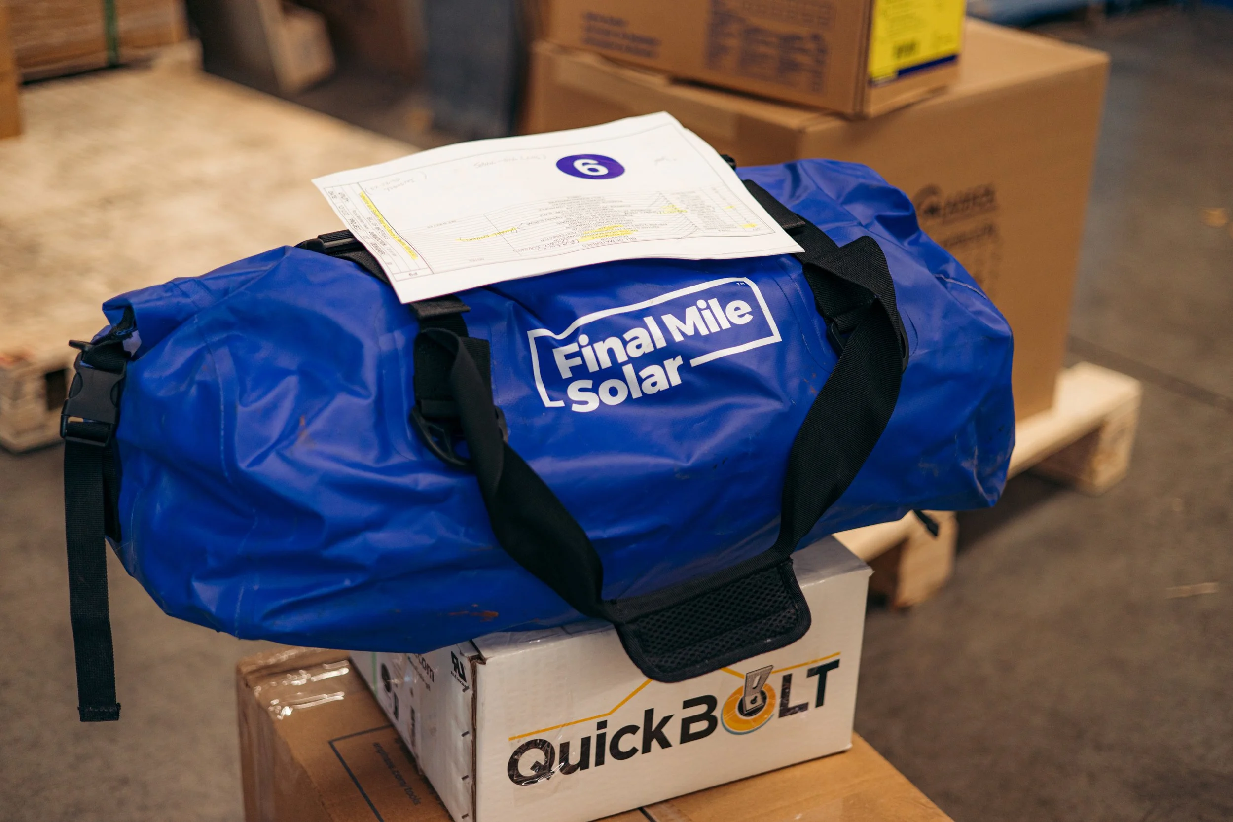 A blue Final Mile Solar duffle bag on a cardboard box labeled QuickBolt, placed on a wooden surface in a warehouse setting with other boxes and pallets visible in the background.