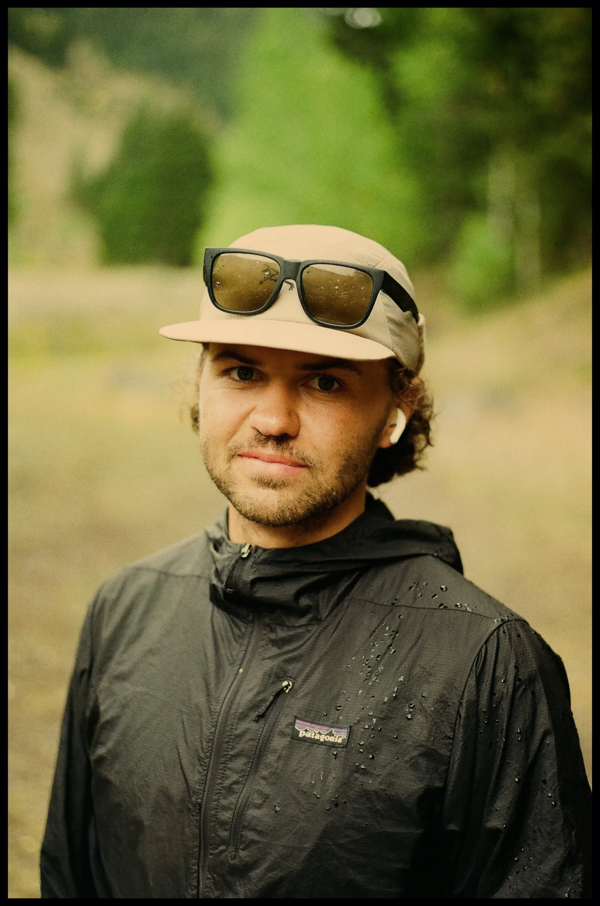 A man with curly hair and a beard outdoors, wearing a beige cap with sunglasses on top, a black wet jacket, and wireless earbuds, standing in a green, blurry natural environment.