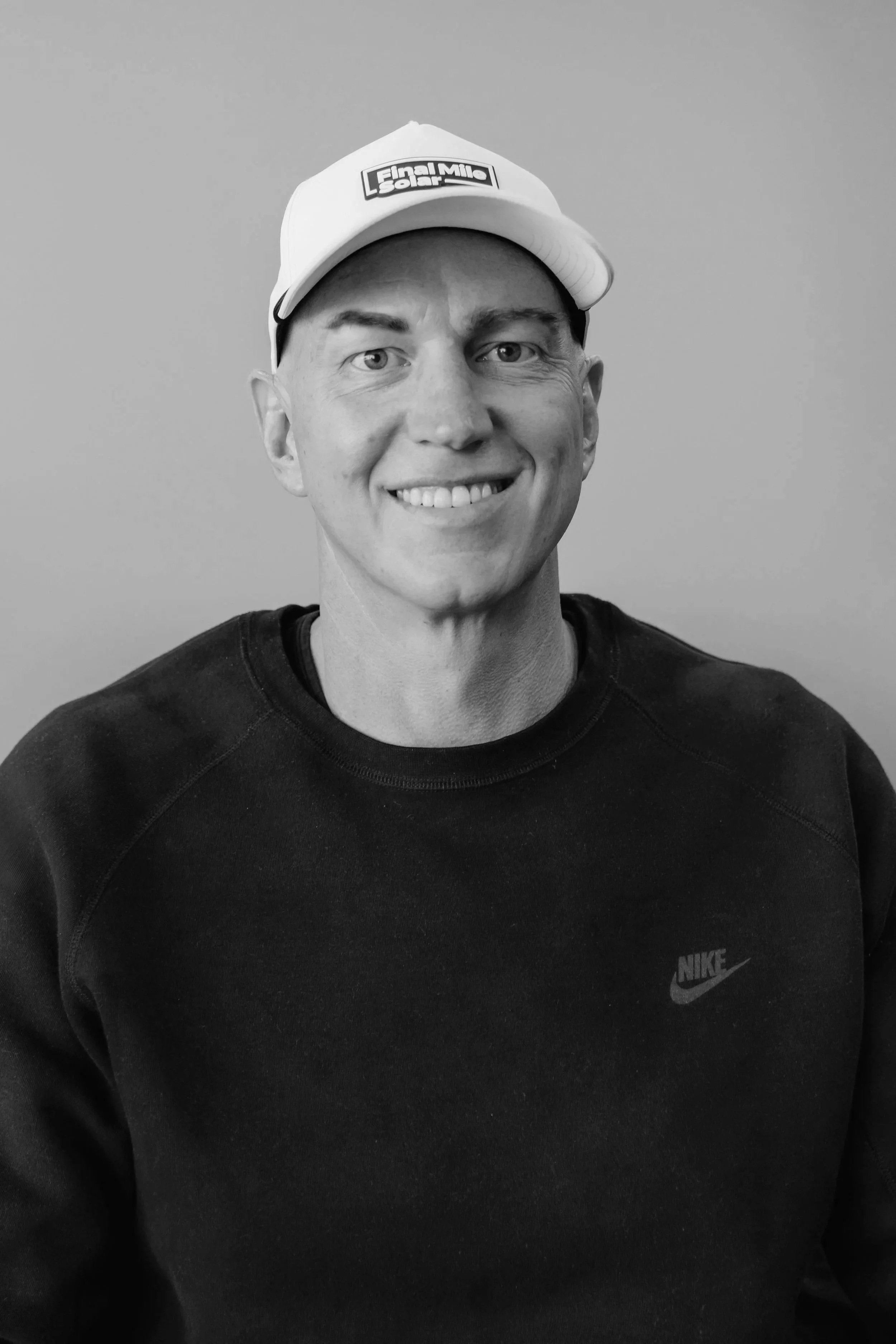 Black and white photo of a smiling man wearing a Nike sweatshirt and a cap that reads "Final Mile Solar."
