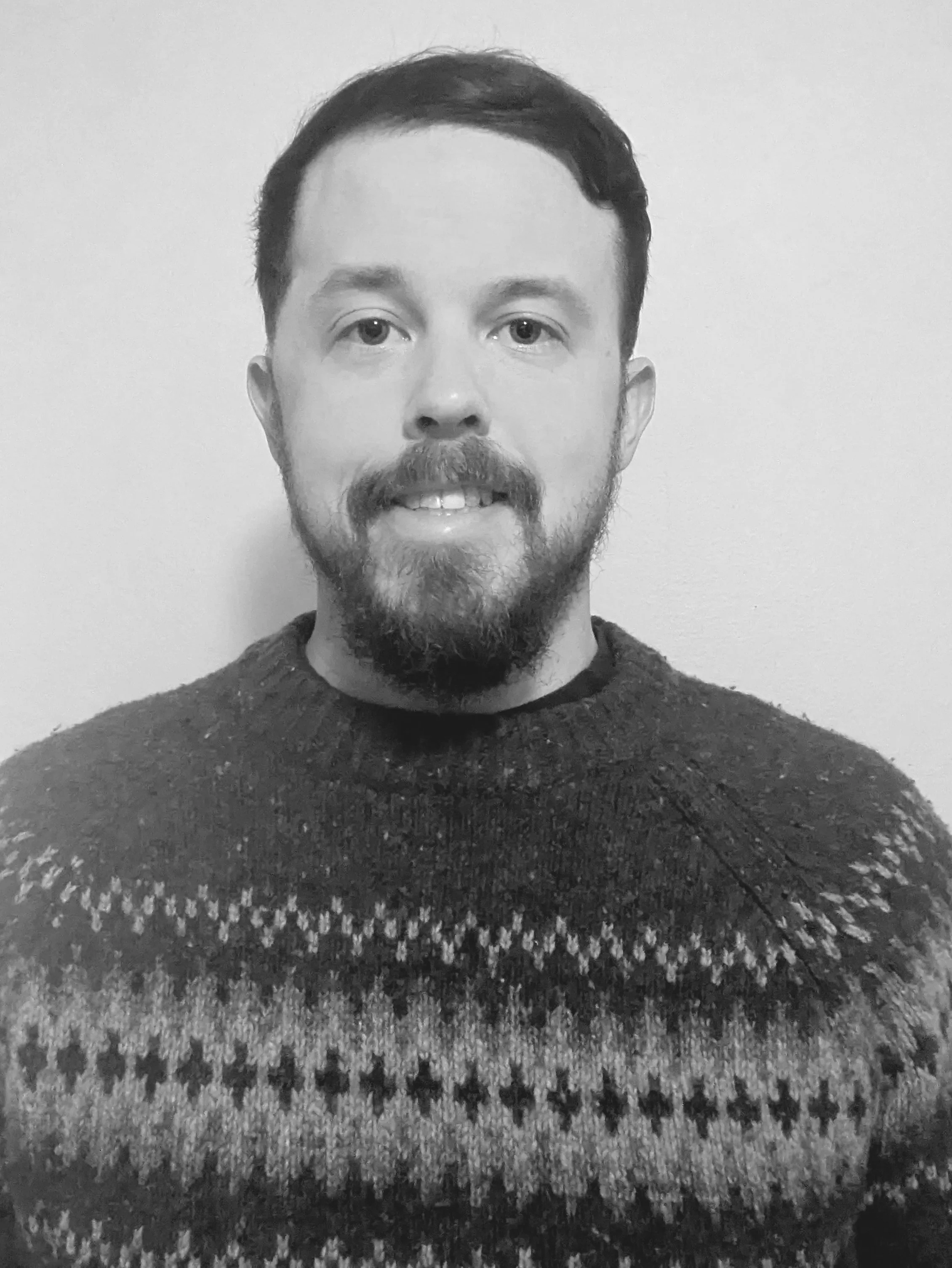 A black and white photo of a man with short dark hair, a beard, and mustache, wearing a sweater with a patterned design, standing against a plain wall.