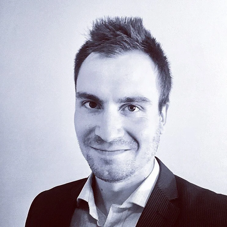 A man with short dark hair, light skin, and a slight smile, wearing a suit jacket over a collared shirt, taking a selfie against a plain background.