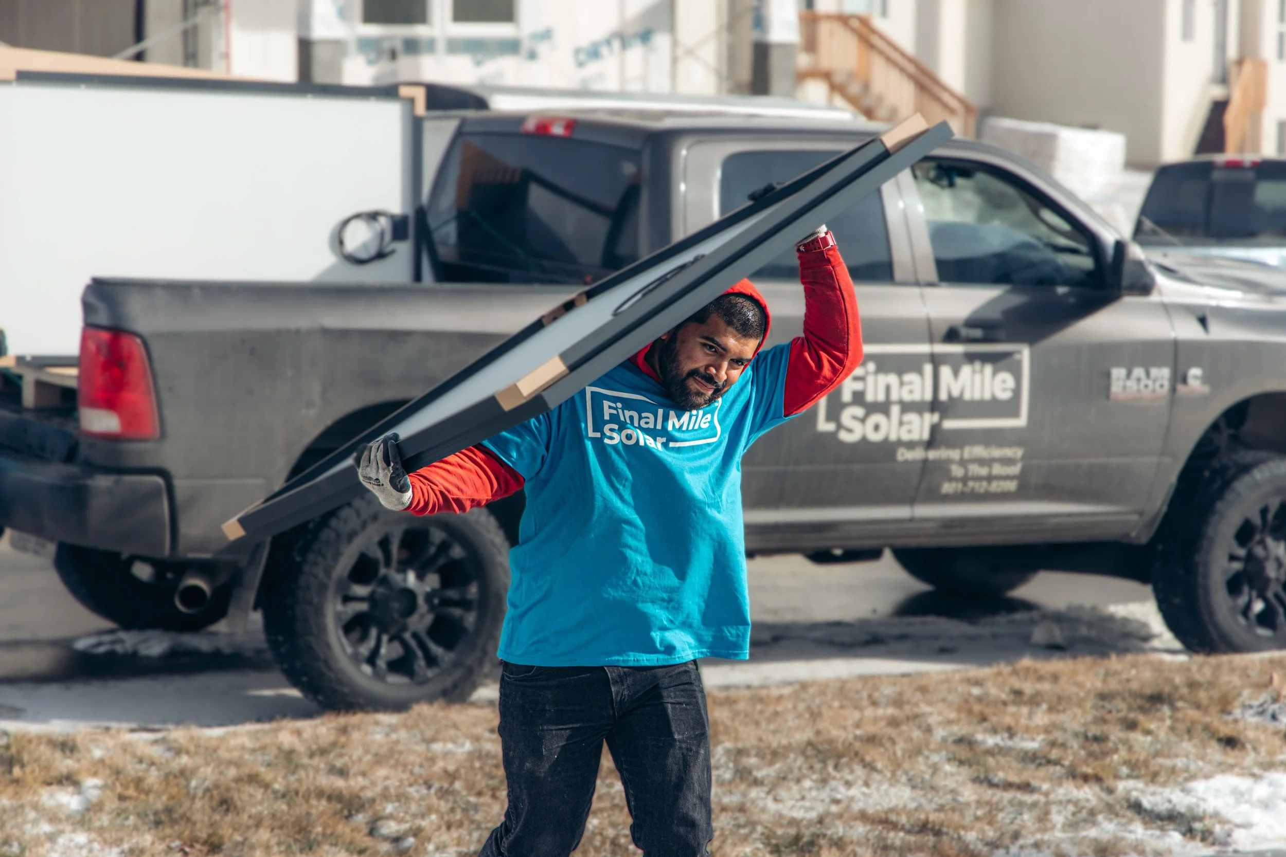 A man is carrying a solar panel on his shoulder in front of a gray pickup truck with 'Final Mile Solar' logo.