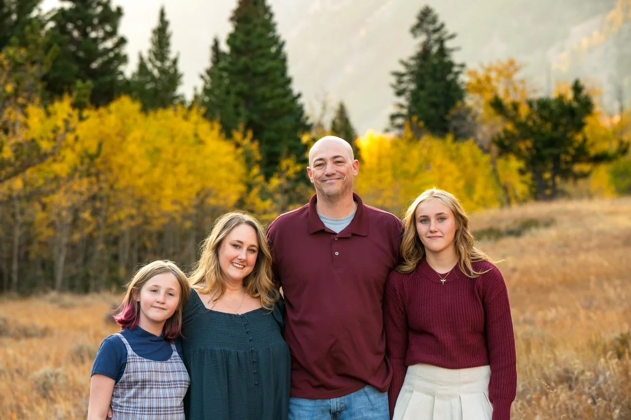 A family of four standing outdoors in a field with autumn trees in the background. The family includes a man, woman, and two girls, all smiling and dressed casually.