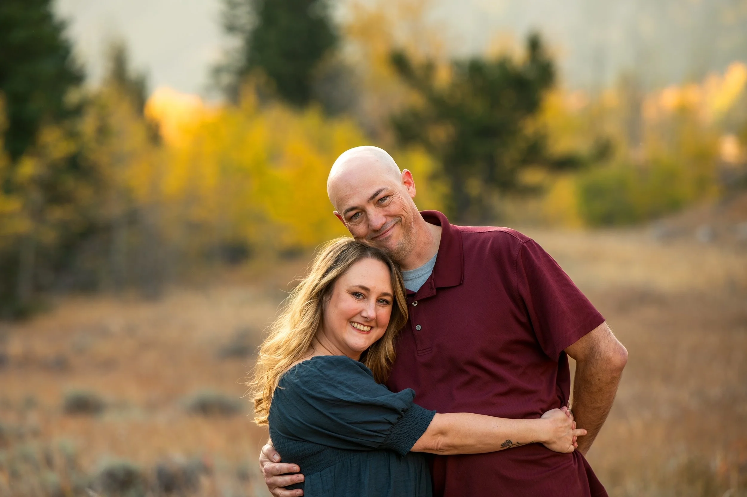 A smiling couple hugging outdoors with trees and autumn leaves in the background.